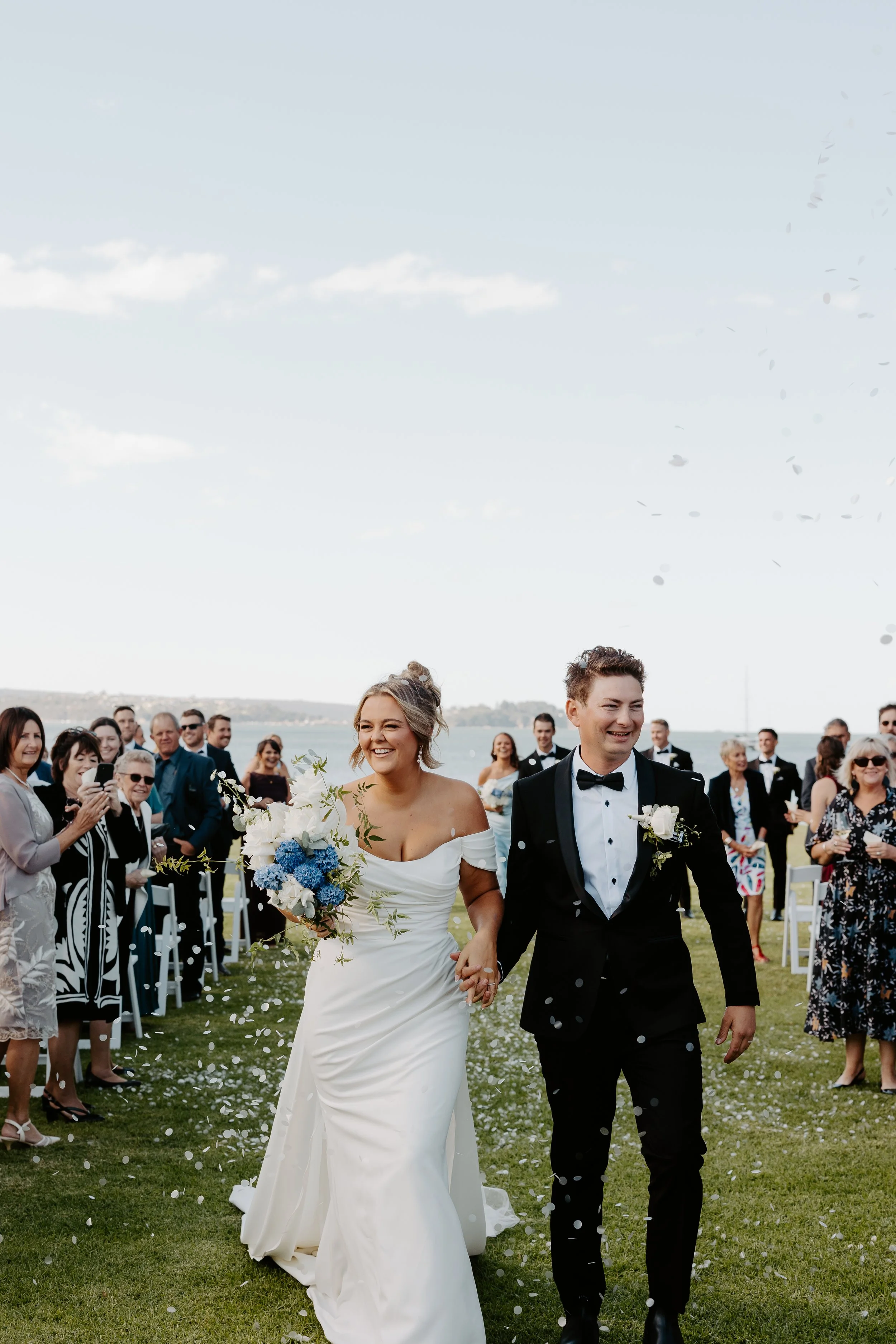 A wedding couple, a woman in a white wedding dress holding a bouquet, and a man in a black tuxedo with a bow tie, happily walking hand-in-hand outdoors on a grassy area with friends and family celebrating, under a partly cloudy sky near a body of wat