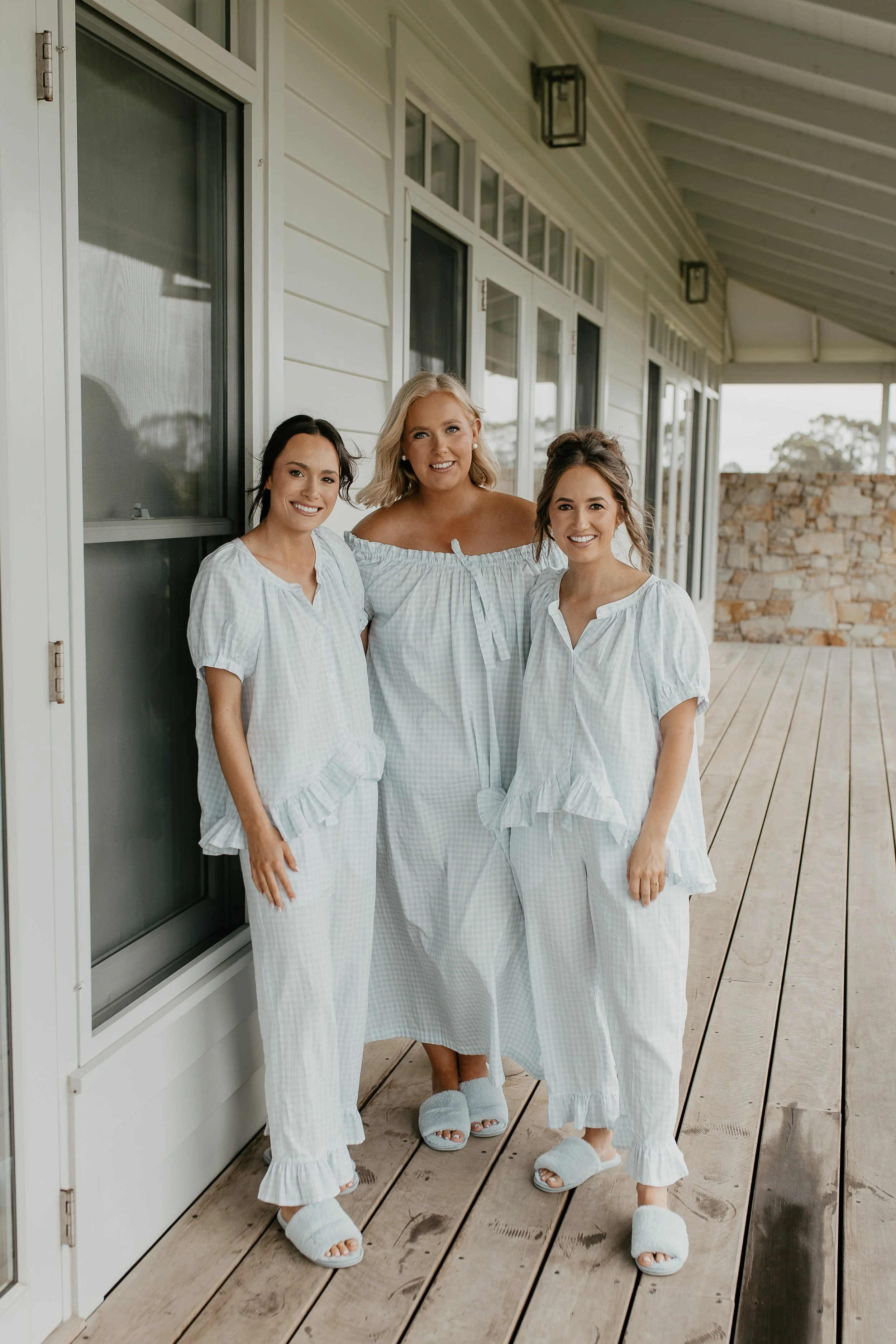 Three women standing on a wooden porch, dressed in white pajamas and slippers, smiling at the camera.
