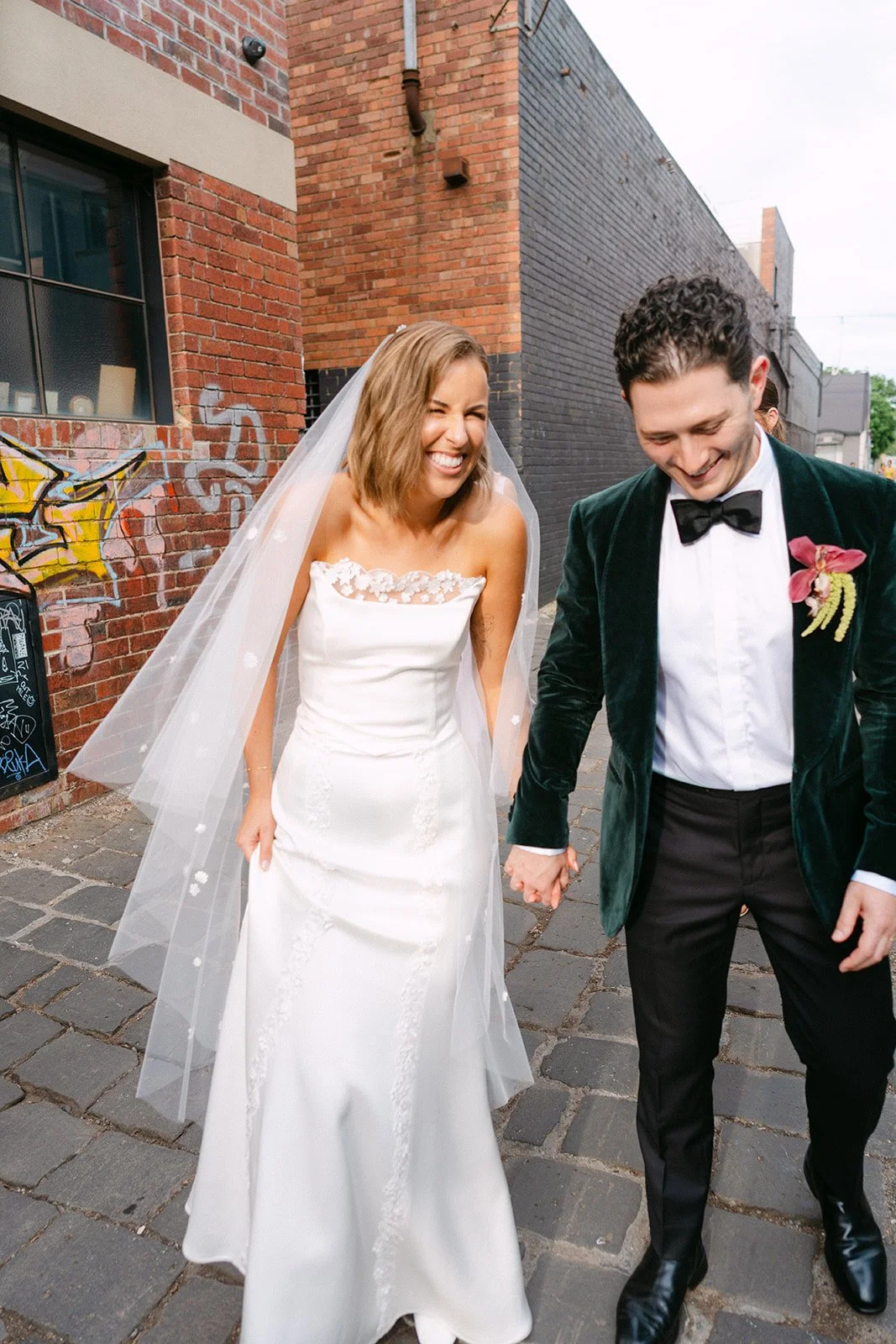 A happy bride and groom holding hands and walking together outdoors, smiling and laughing. The bride is wearing a white wedding gown and veil, and the groom is dressed in a dark green velvet jacket with a black bow tie. They are on a cobblestone stre