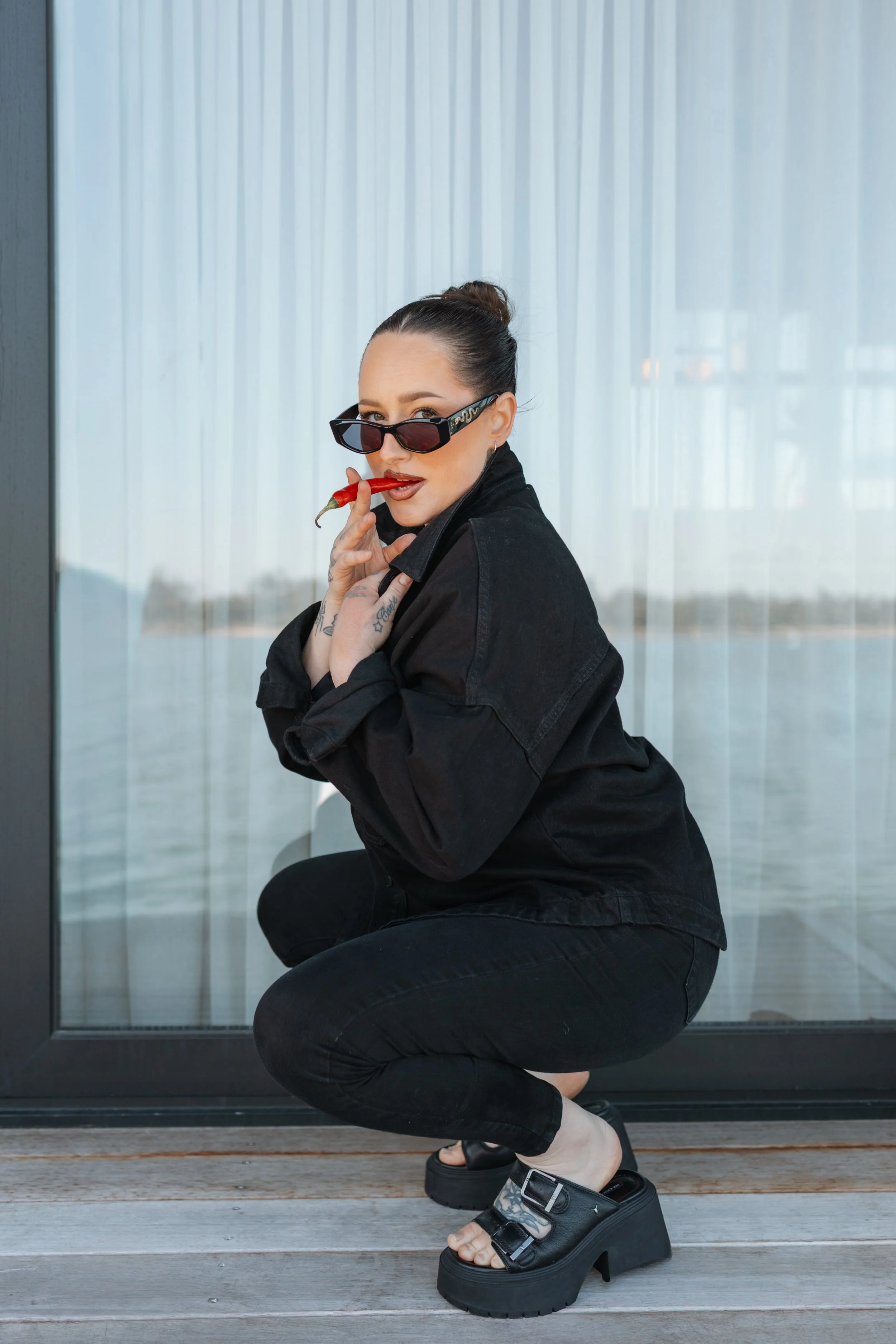 A woman crouching in front of a window with sheer curtains, wearing black sunglasses, a black jacket, and black platform sandals with thick heels, holding a red chili pepper in her mouth and touching her face with her left hand.