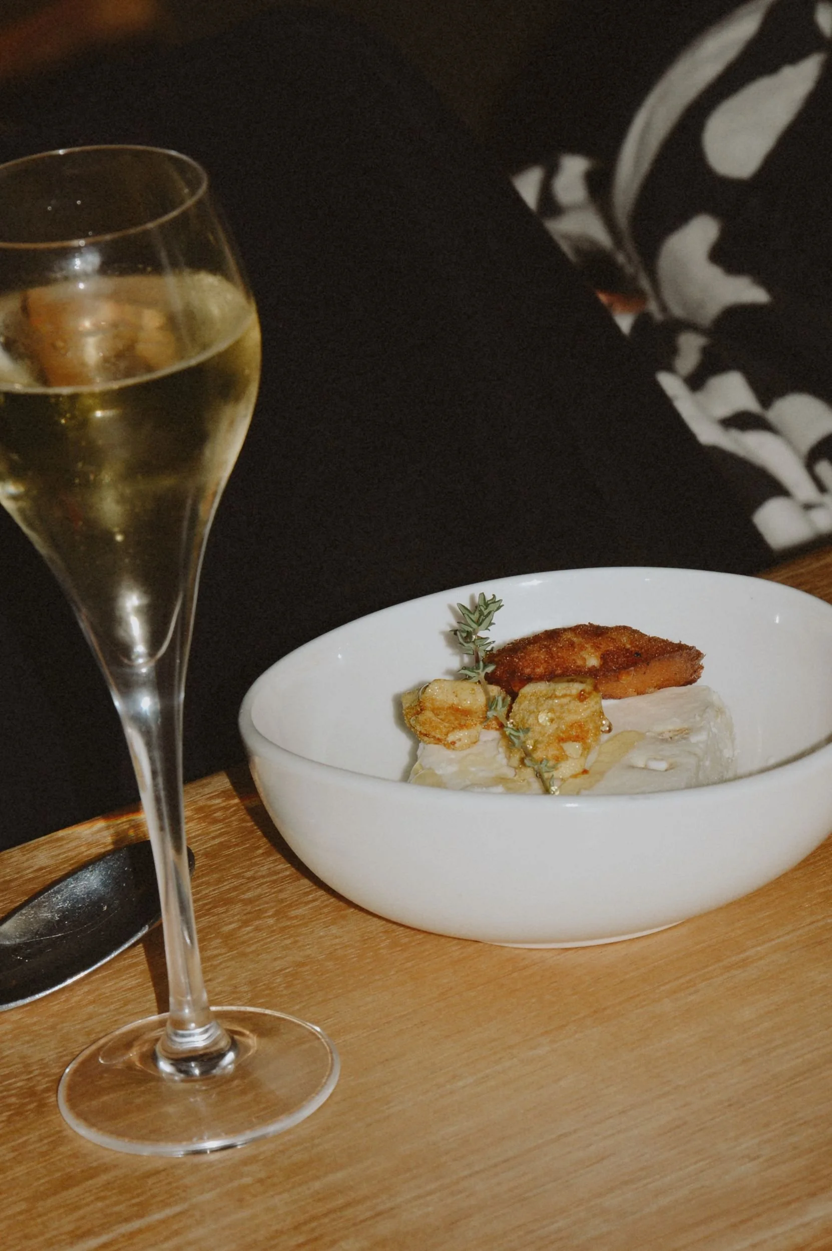 A glass of white wine and a bowl of food on a wooden table, with a person wearing a black and white patterned shirt partially visible in the background.