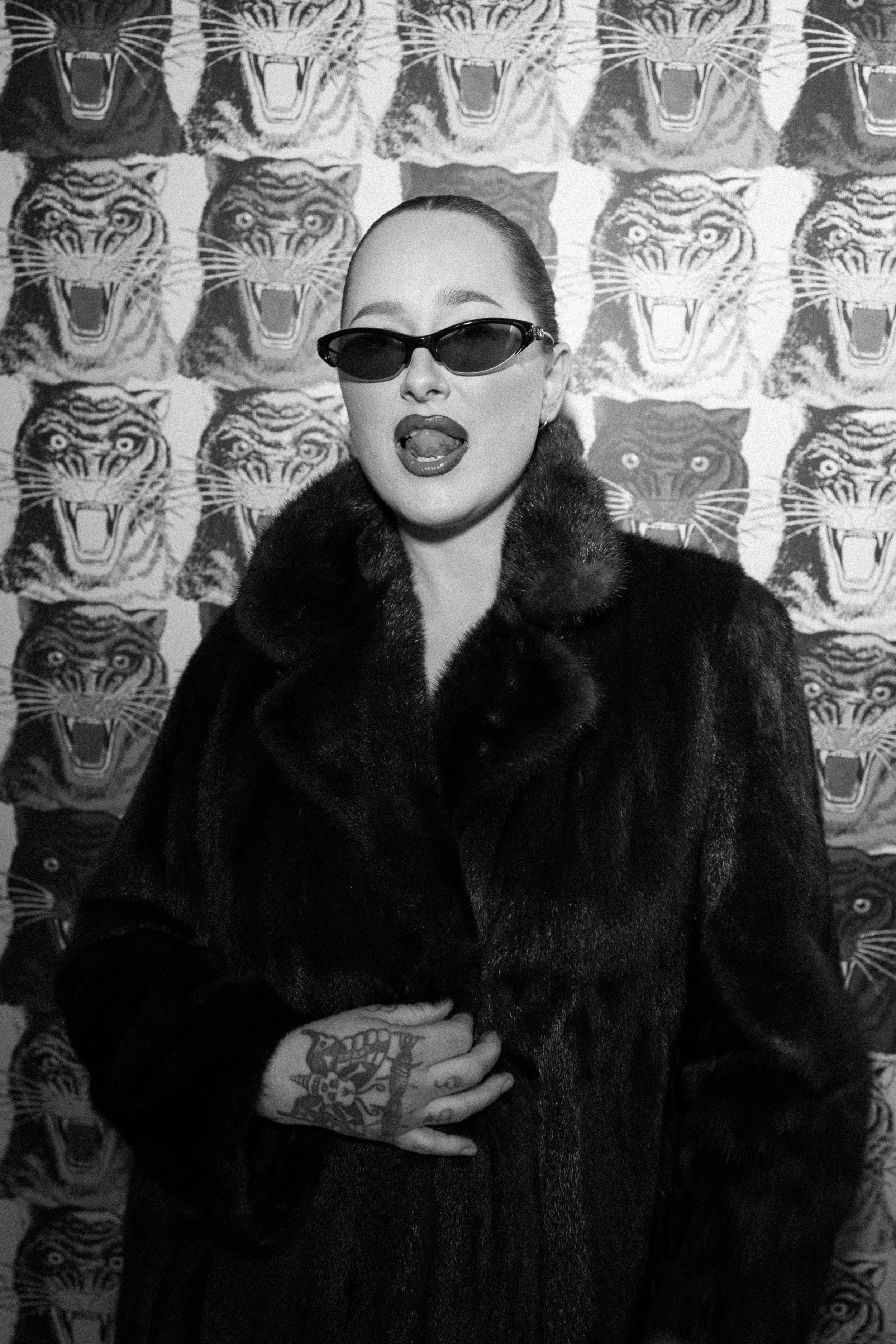 A woman in a fur coat and sunglasses sticking out her tongue on a backdrop with tiger faces.