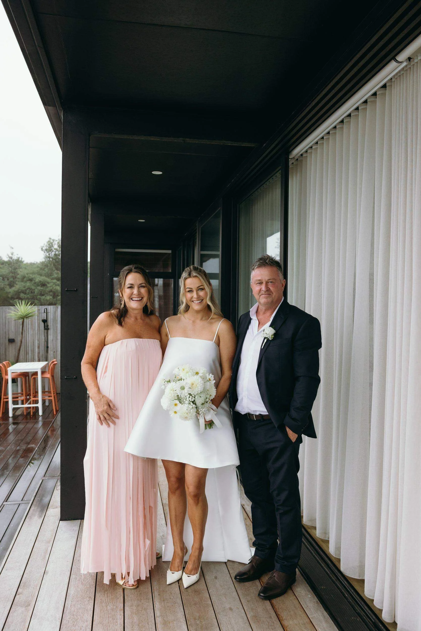 Three people smiling and standing together outdoors on a porch, with two women and one man. The woman in the middle is dressed in a white wedding gown holding a bouquet, and the other woman is wearing a light pink strapless dress. The man is dressed 