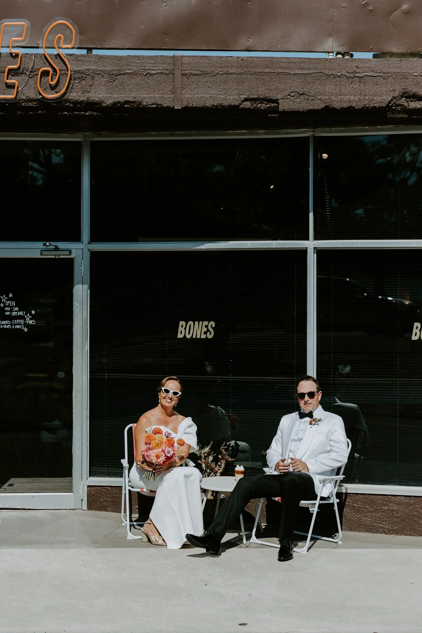 A bride and groom sitting outside in front of a glass storefront with the word "BONES" on it. The bride is holding a bouquet of pink and orange flowers, wearing white sunglasses and a white dress. The groom is wearing a white tuxedo shirt, black pant