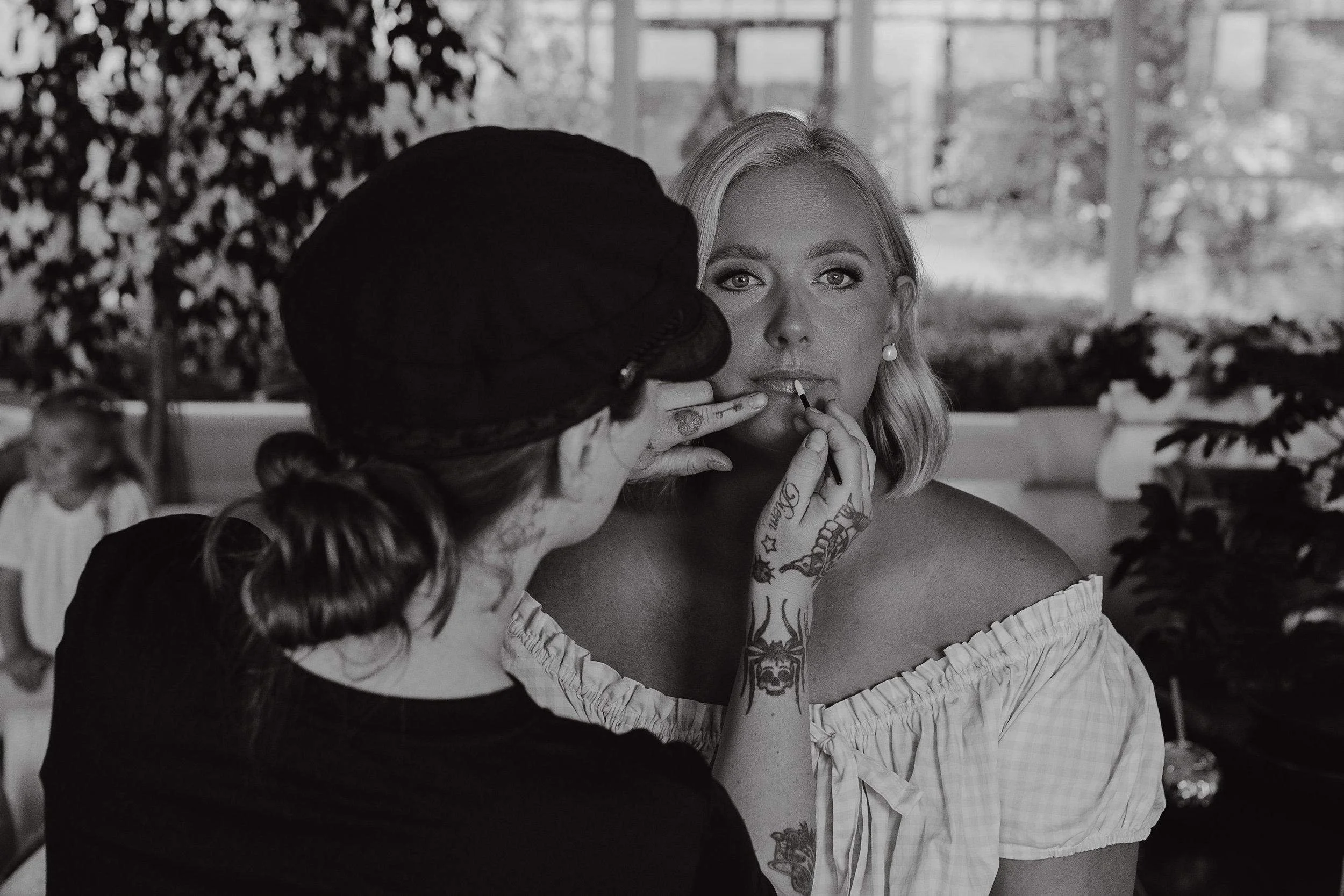 A makeup artist applies lipstick to a woman with light-colored hair, tattoos, and jewelry, in a well-lit room filled with plants and large windows.