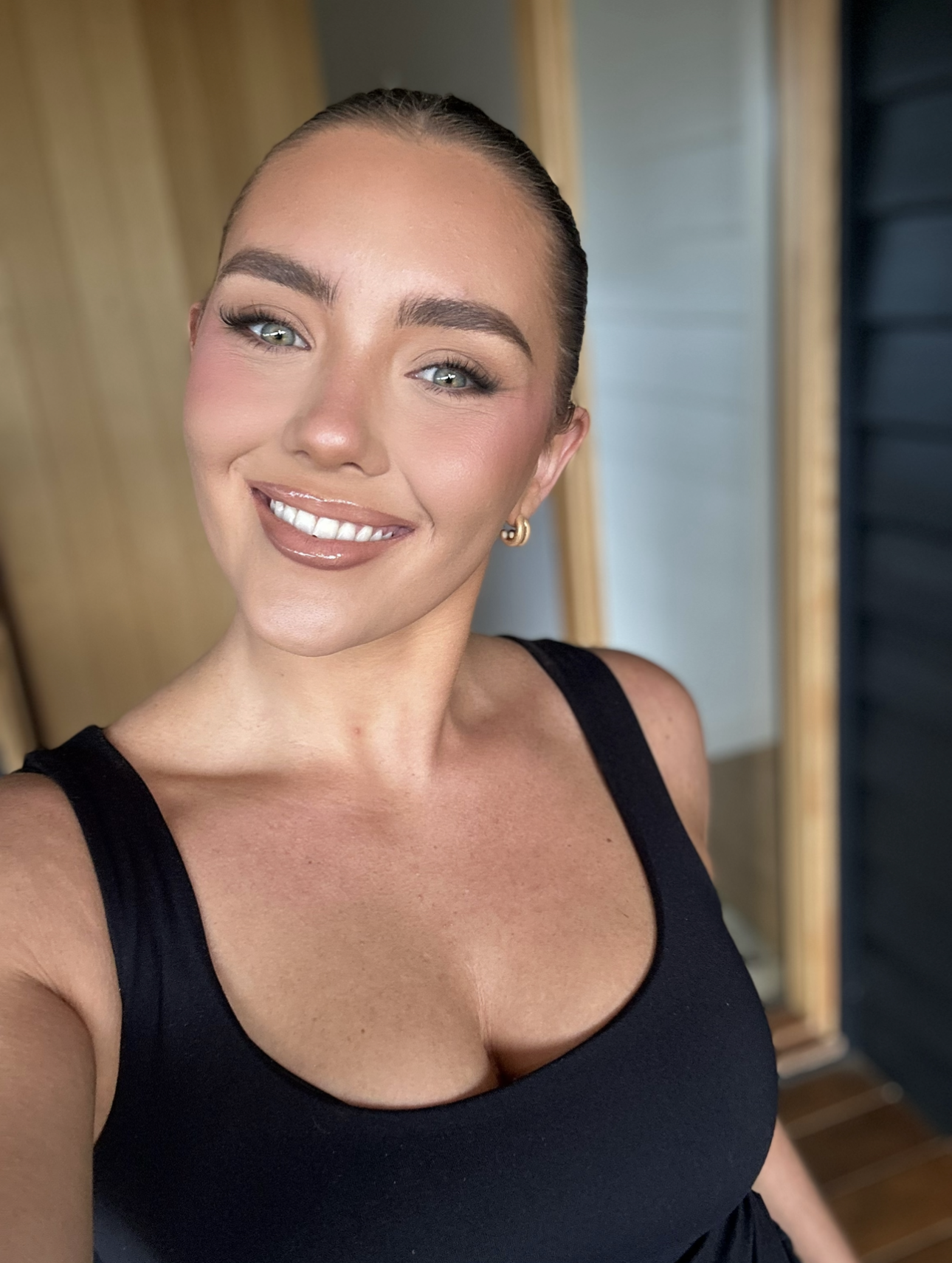 Close-up of a smiling woman with light makeup, wearing a black tank top, gold hoop earrings, and her hair pulled back in a sleek style.