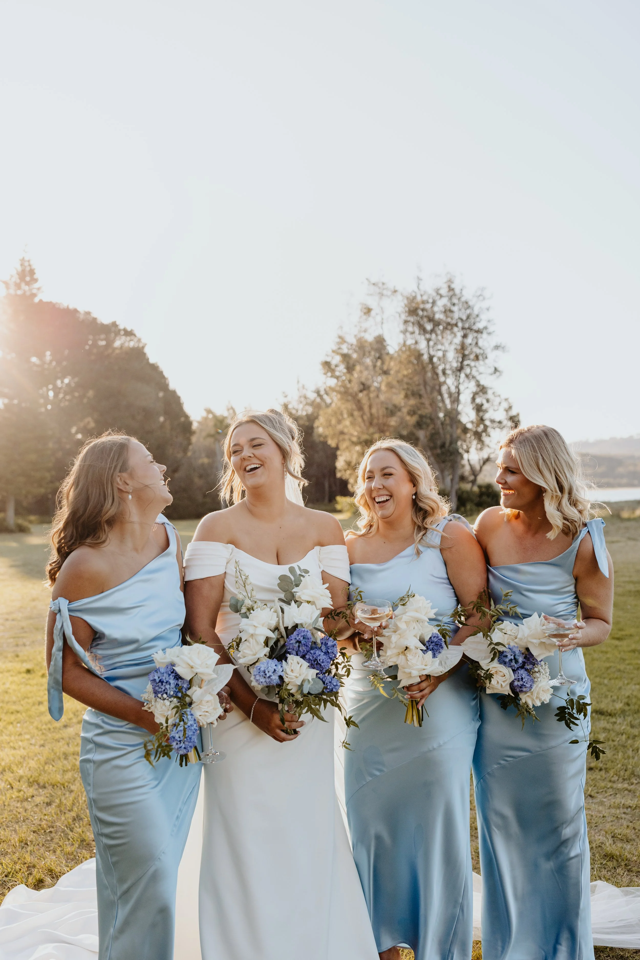 A bride with four bridesmaids standing on a lawn during a wedding celebration in the late afternoon or early evening, all holding bouquets with white and purple flowers.