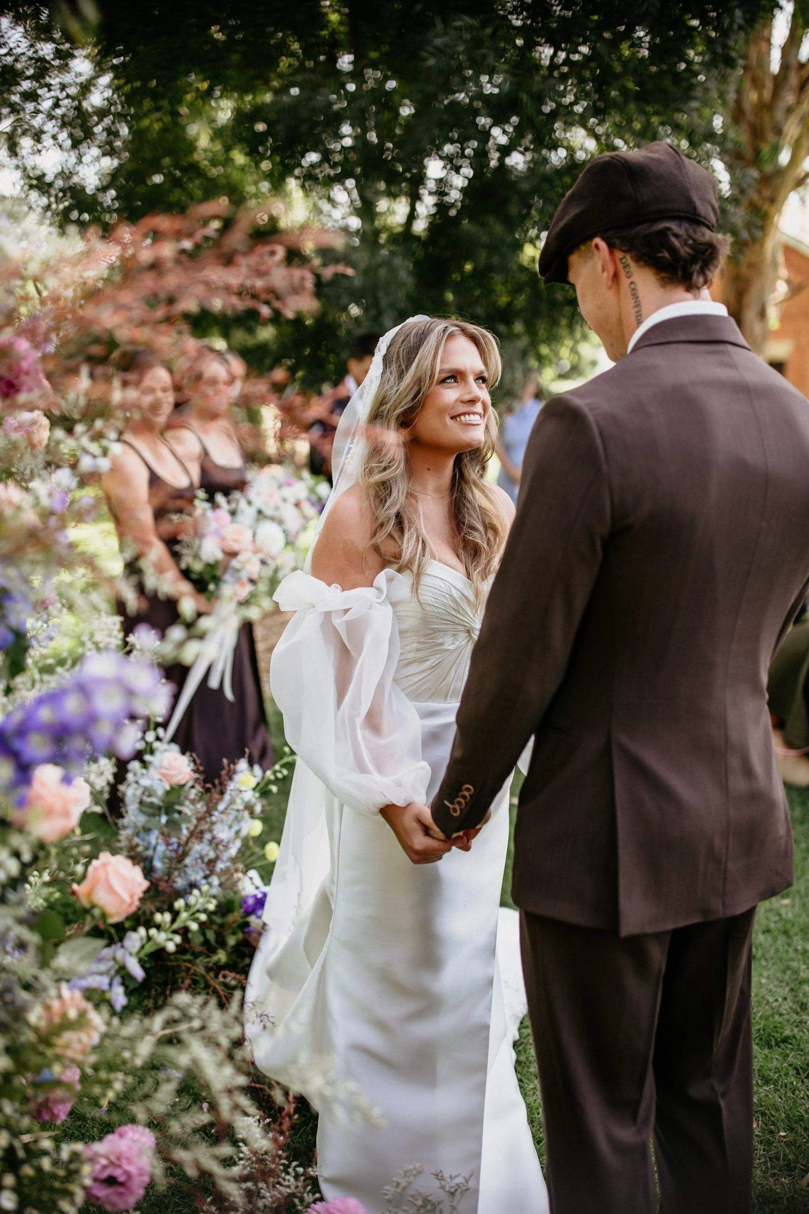 A bride and groom holding hands during a wedding ceremony outdoors, with bridesmaids and floral arrangements in the background.