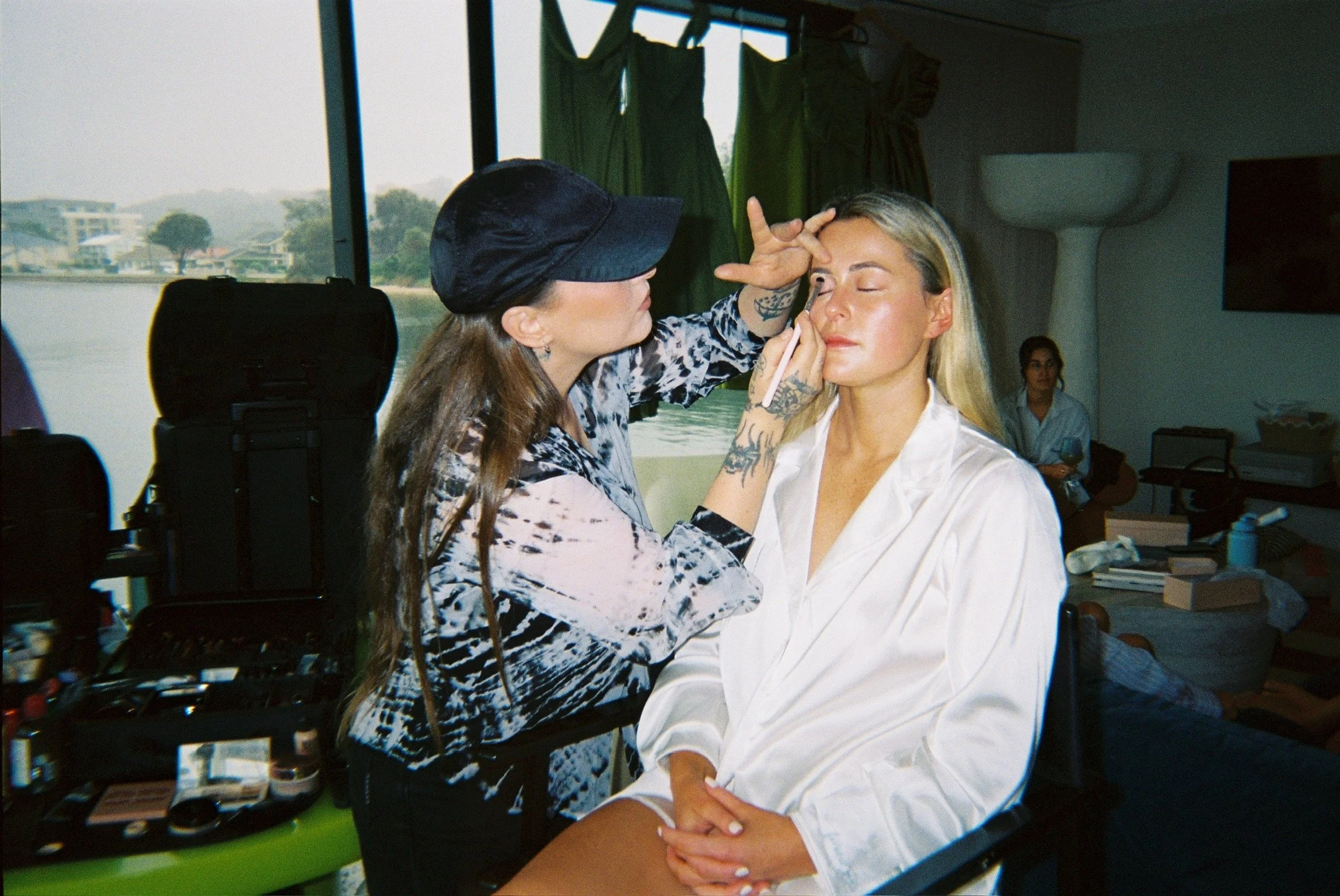 A makeup artist applies makeup to a woman seated with her eyes closed in a room with large windows and green curtains.