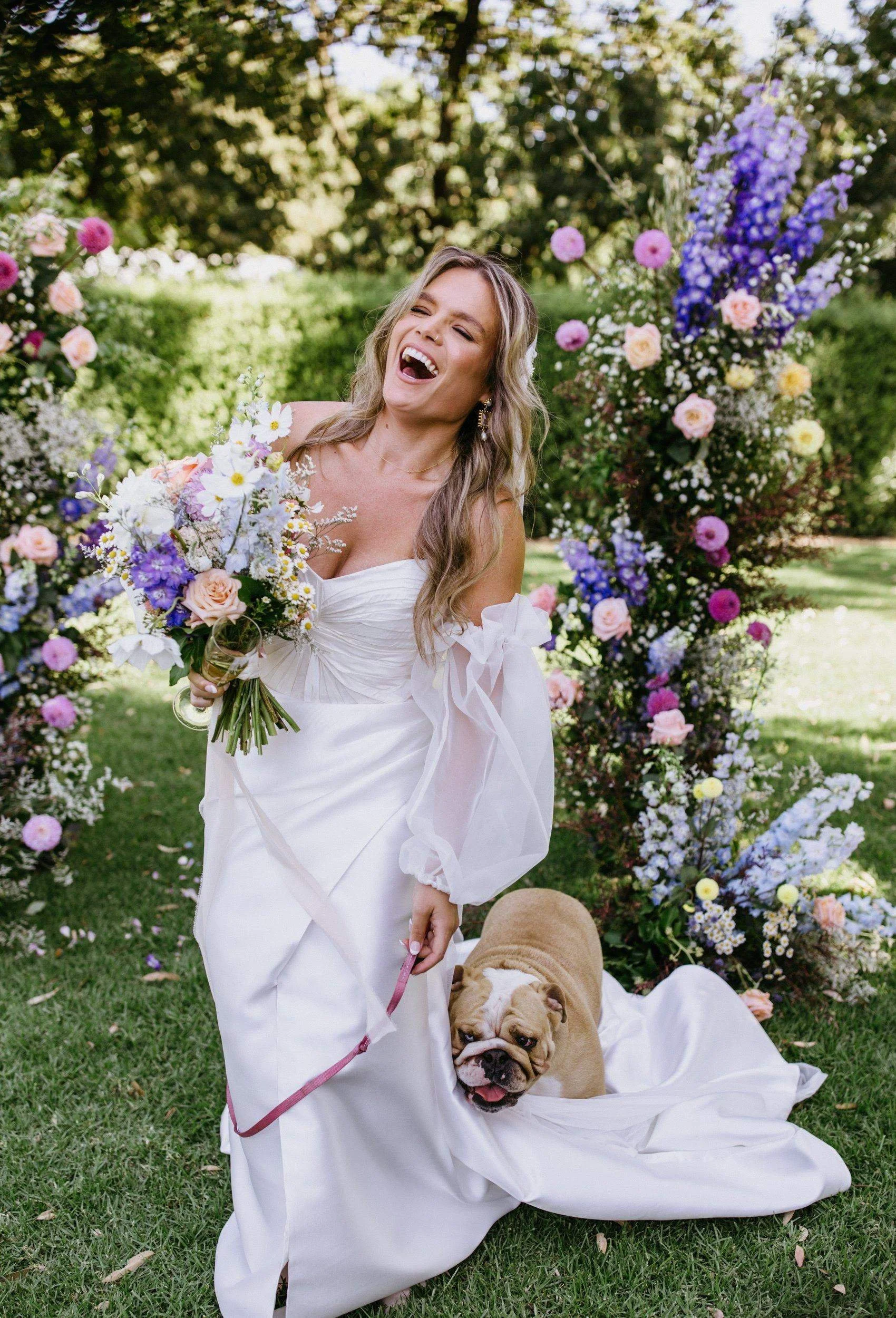 A woman in a white dress holding a bouquet of flowers, smiling and laughing outdoors with a garden of colorful flowers and greenery in the background. A bulldog puppy is sitting in a white cloth on the ground.
