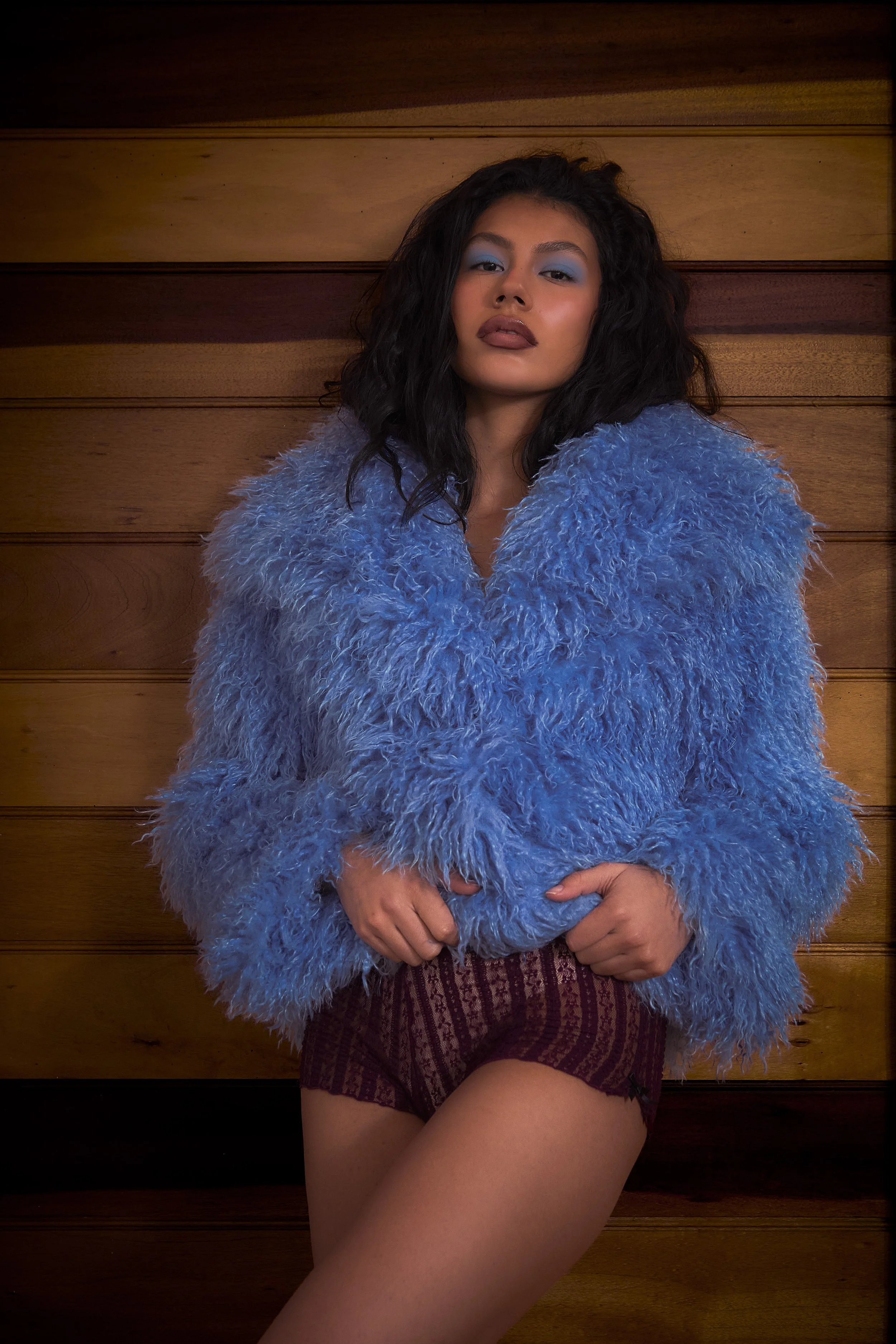 A woman with curly black hair is wearing a light blue fuzzy coat and patterned shorts, standing against a wooden wall background.