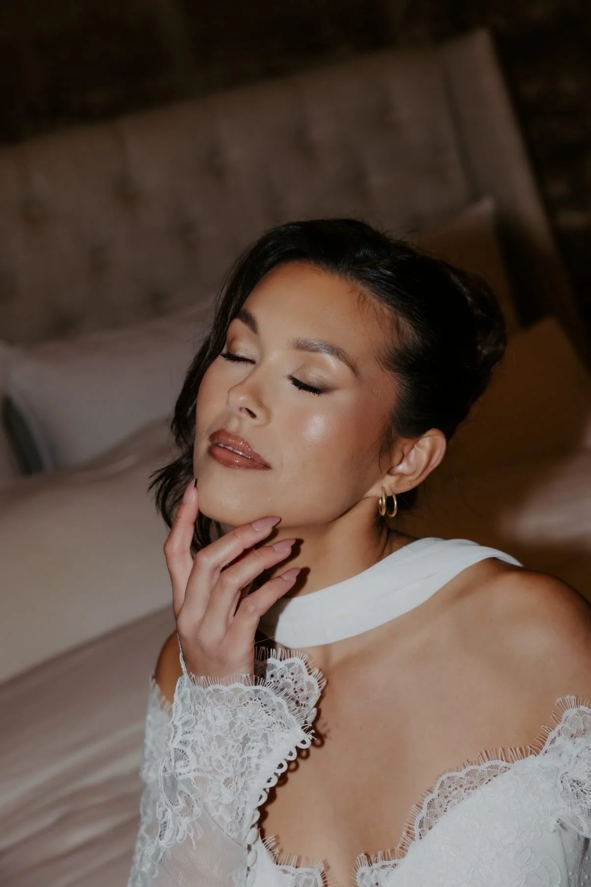 A woman with dark hair and earrings resting her chin on her hand, life her eyes closed, in a white lace top sitting on a bed with a tufted headboard.