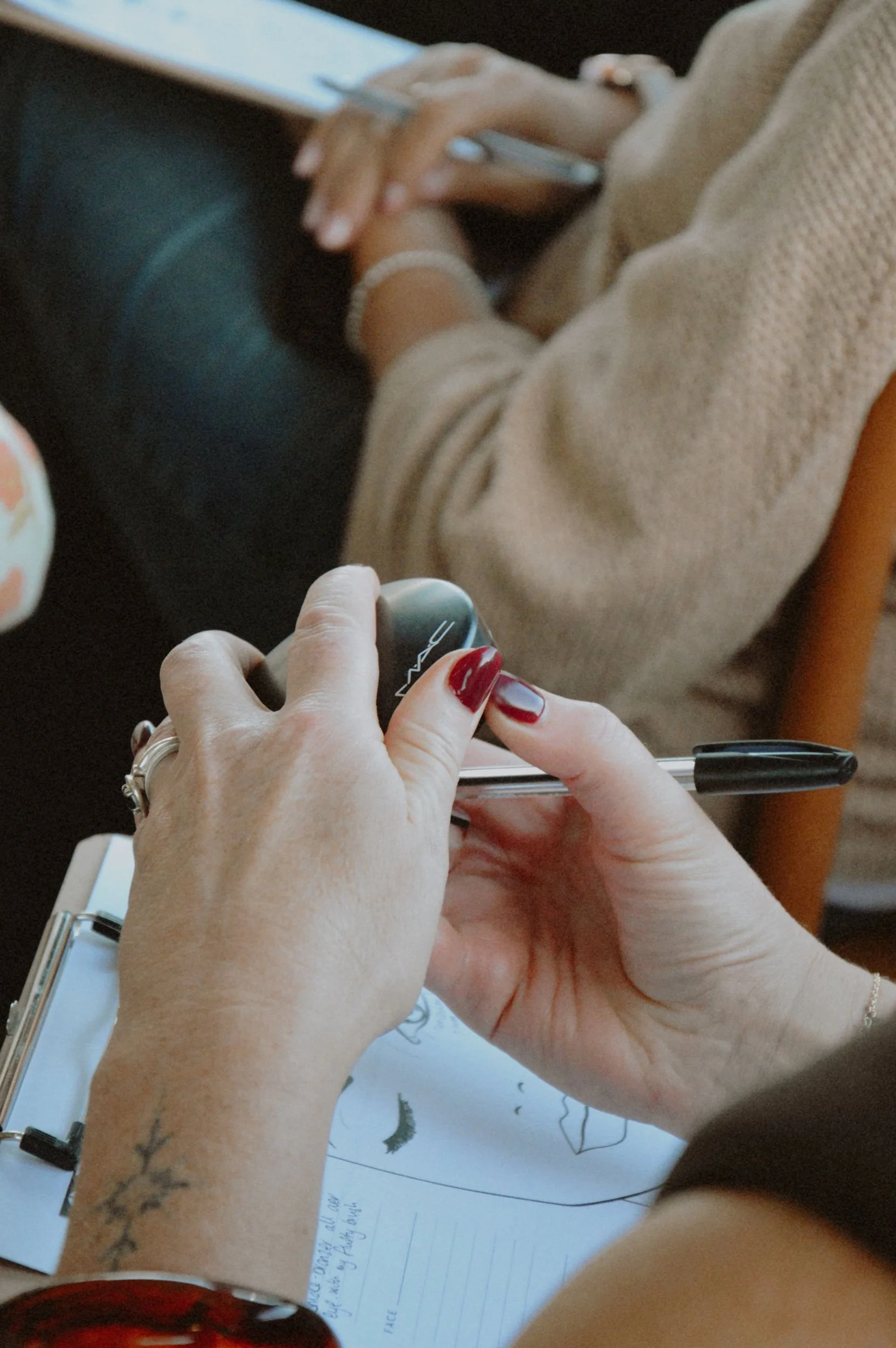 Multiple hands are engaged in drawing on a piece of paper, using various pens and markers. One person has a tattoo on their arm and is wearing a bracelet and rings. The scene appears to be a creative or artistic activity.