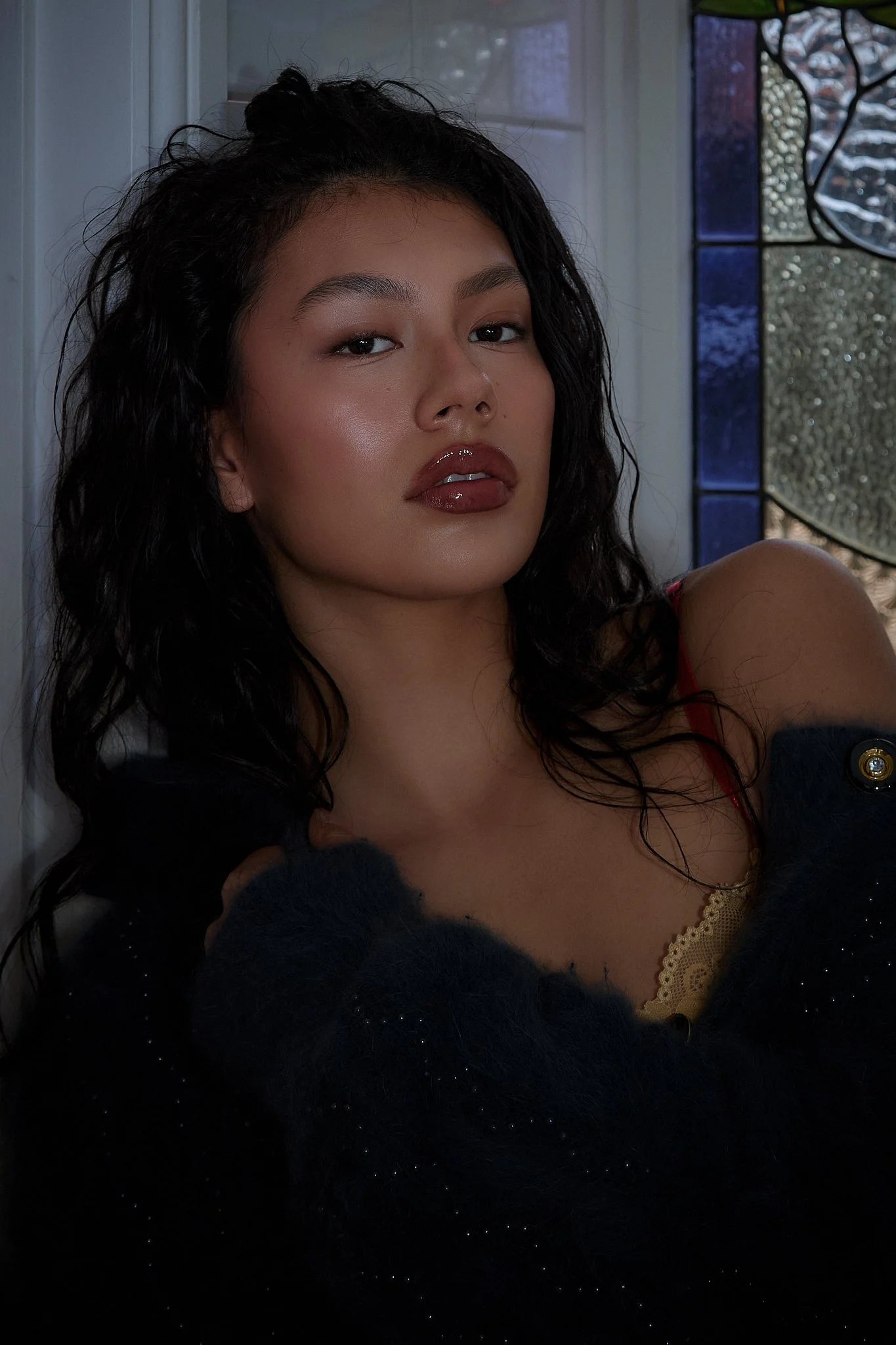 A young woman with dark, wavy hair styled in a half updo, wearing glossy lipstick and eye makeup, posing near a window with colorful stained glass. She is dressed in a yellow lace top with a black fuzzy cardigan, with a neutral expression.
