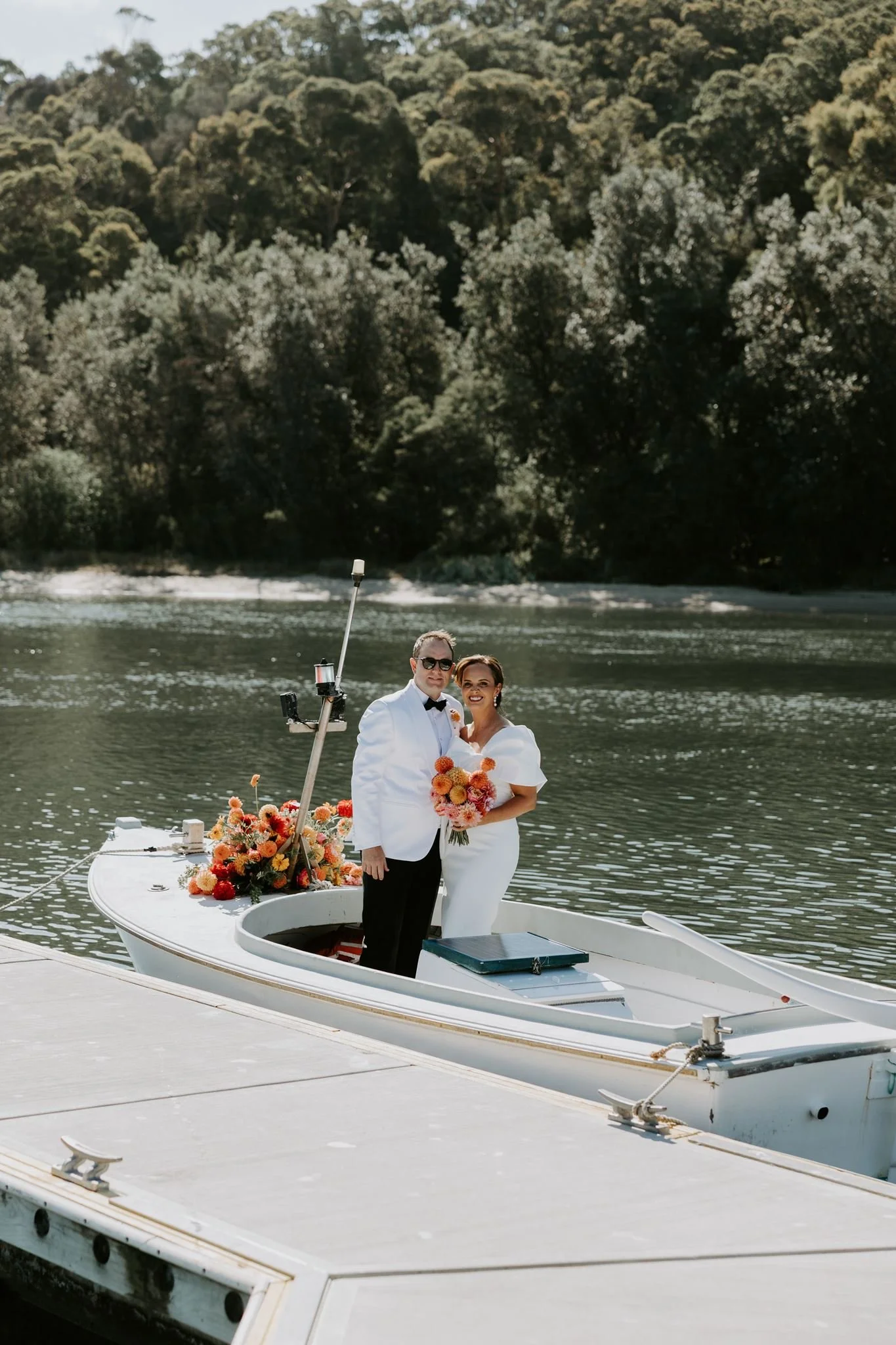 A bride and groom standing on a boat decorated with flowers on a body of water, with a background of green trees.