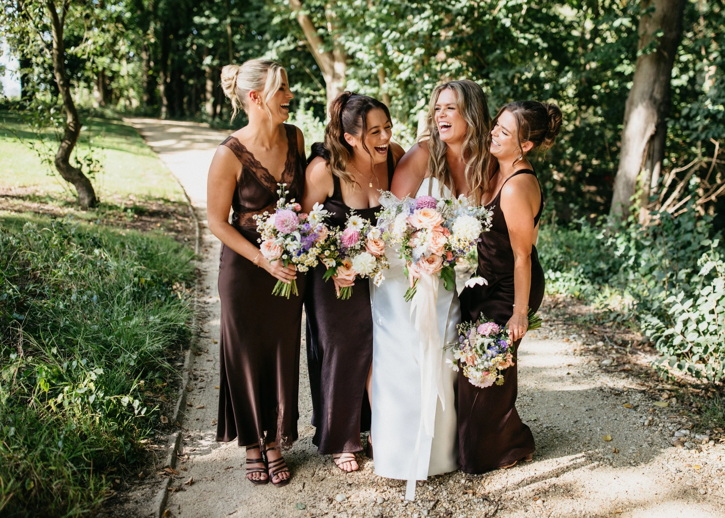 Four women dressed in formal attire walking outdoors on a dirt path, holding bouquets of flowers and laughing together under shaded trees.