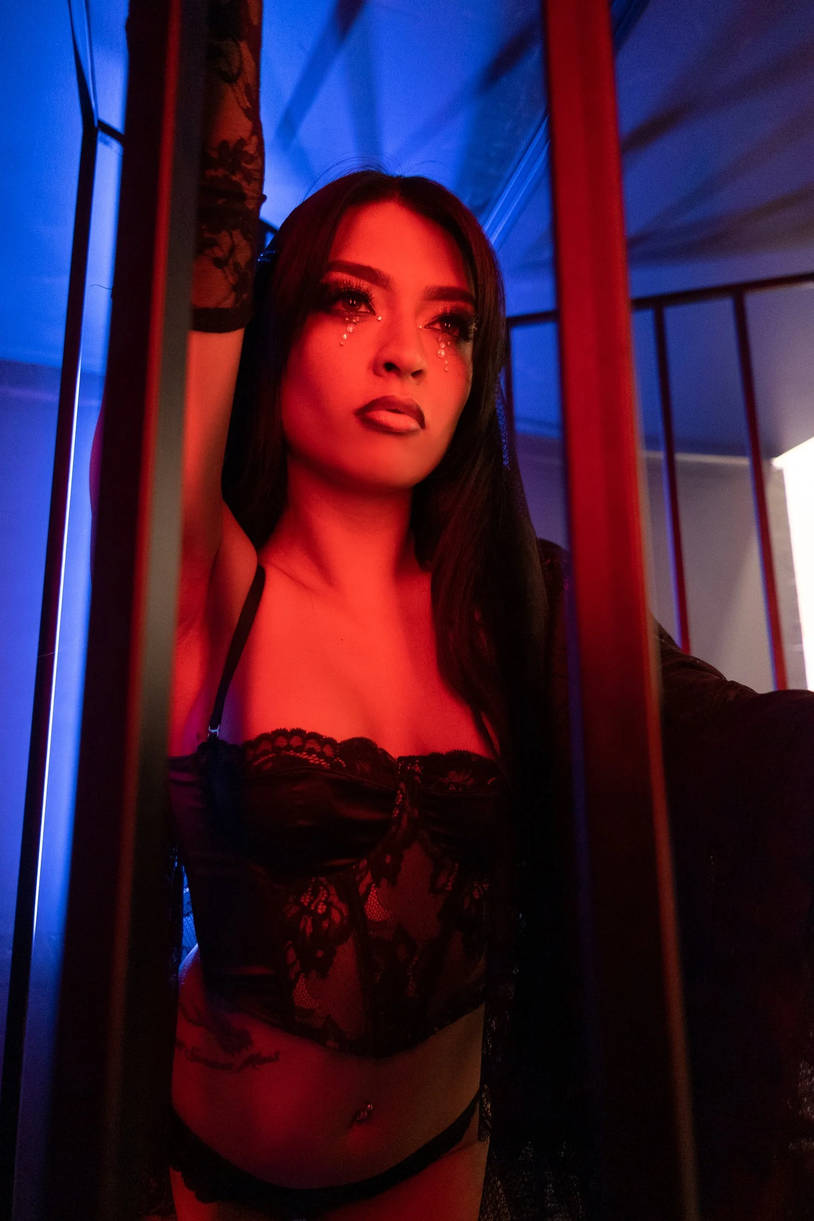 Woman in black lace lingerie with glitter makeup, posing behind bars with colorful blue and red lighting.
