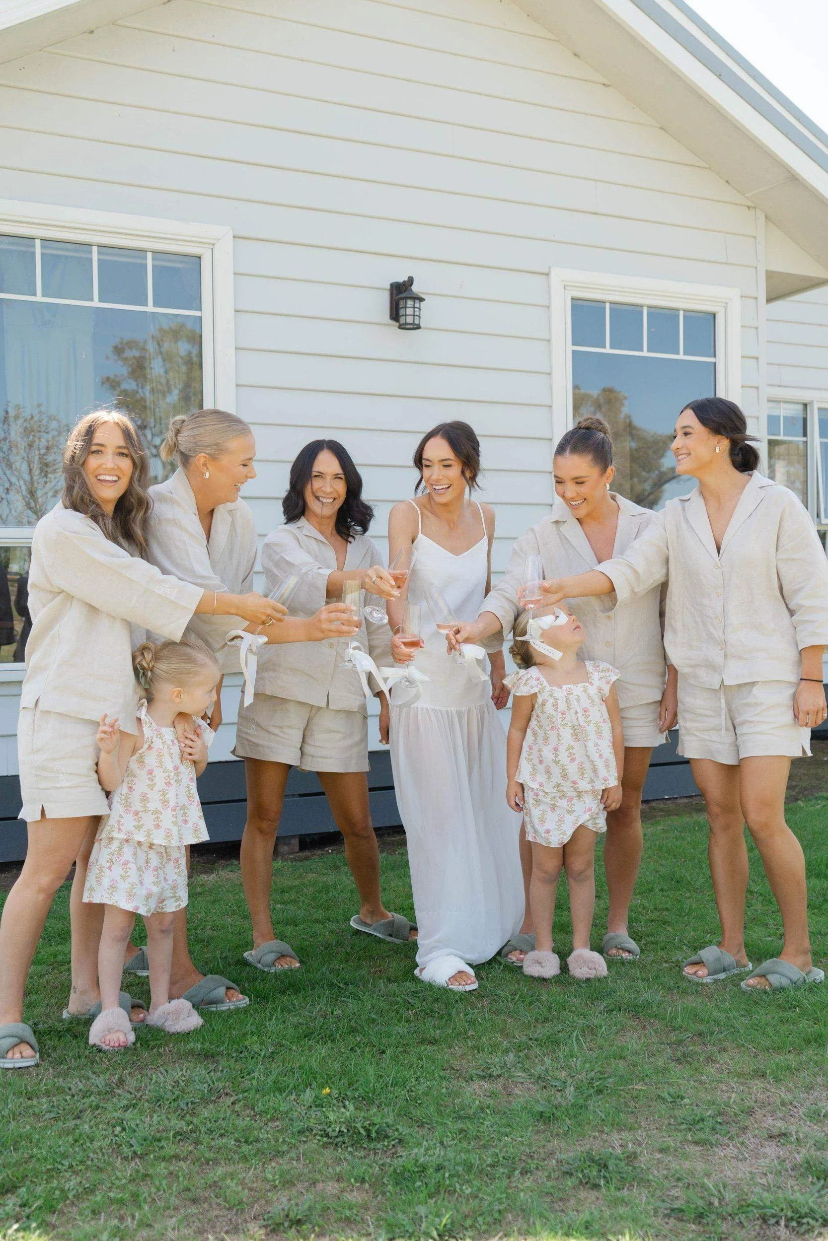 Group of women and two young girls standing outside on grass, dressed in matching beige pajamas, smiling and holding glasses of pink wine, in front of a white house with large windows.