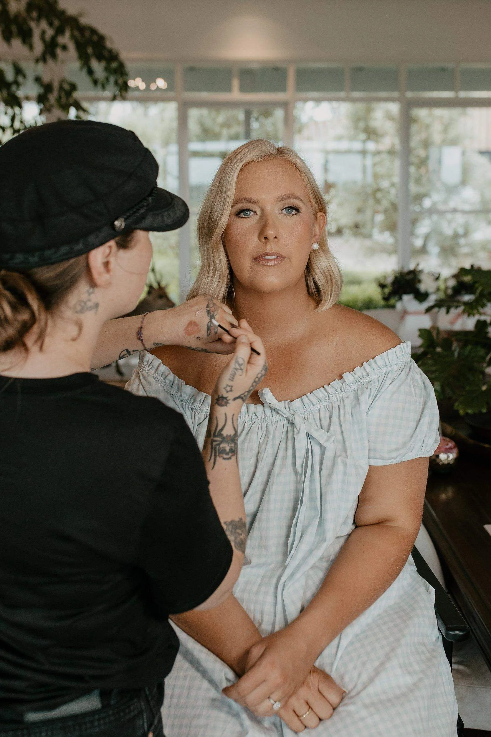 A woman with blonde hair sitting in a room, getting her makeup done by another person wearing a black cap and shirt.