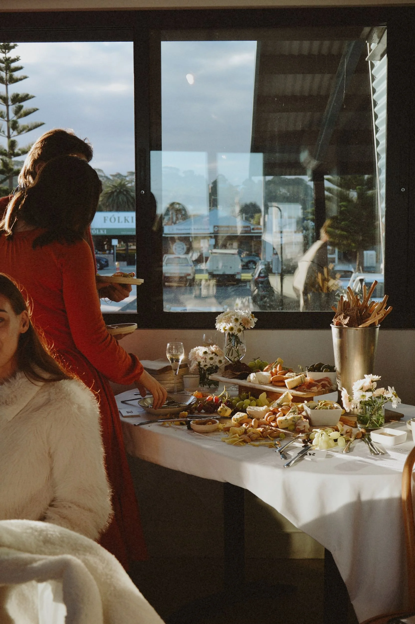 Interior of a restaurant with a table set with a cheese and charcuterie platter, flowers, and glasses. Two people are standing near the table, one holding a plate. Through the large window, a parking lot and trees are visible outside.