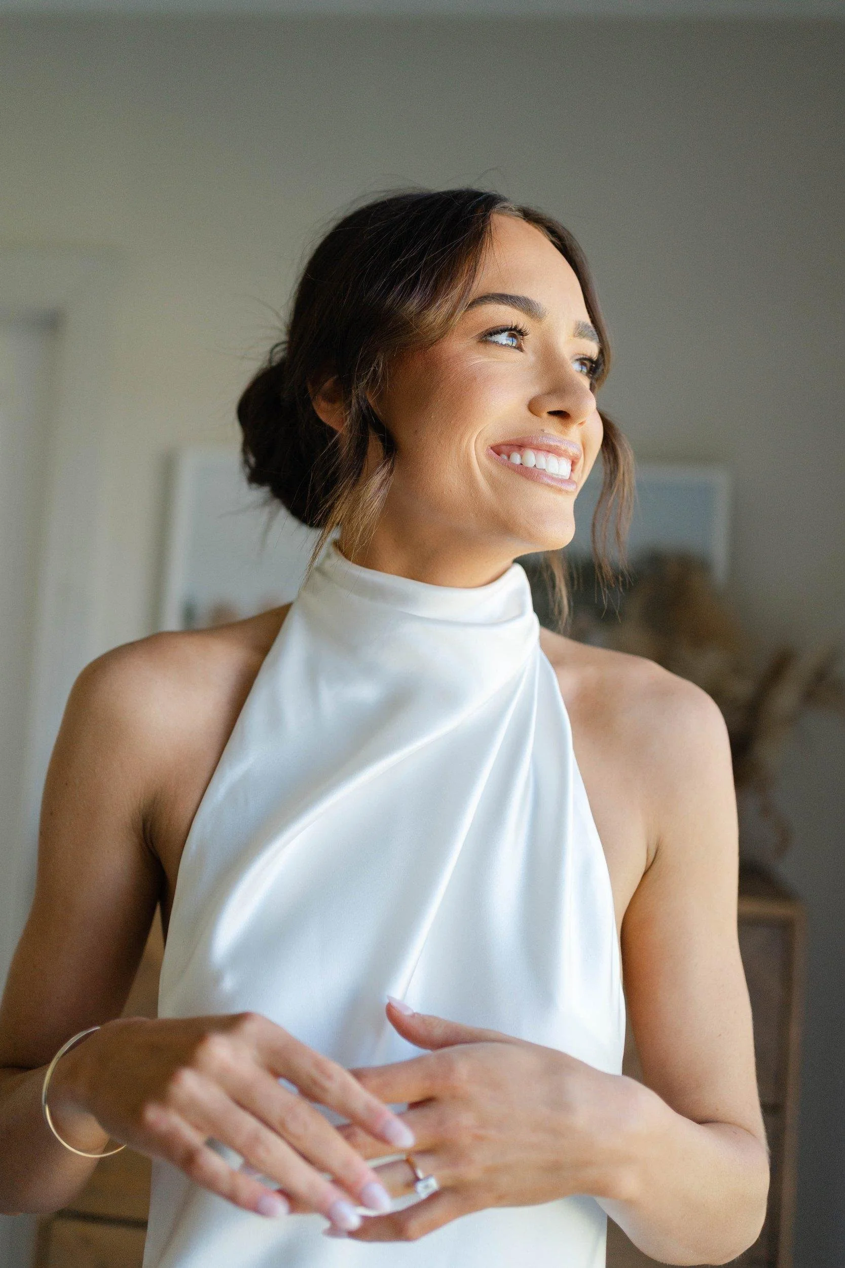 A woman with short brown hair styled in a messy bun, wearing a white sleeveless satin gown, smiling and looking to her right, standing indoors.