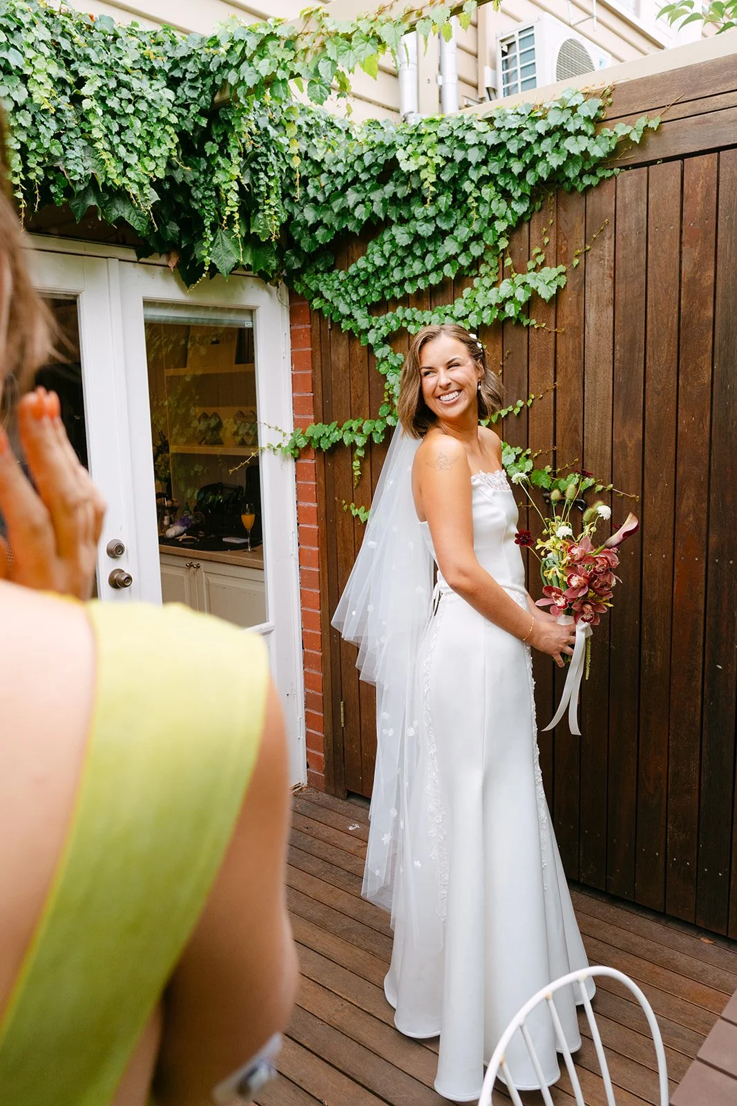 A bride in a white wedding dress holding a bouquet, smiling and standing against a wooden fence. Another person, partially visible, appears to be reacting in front of her.