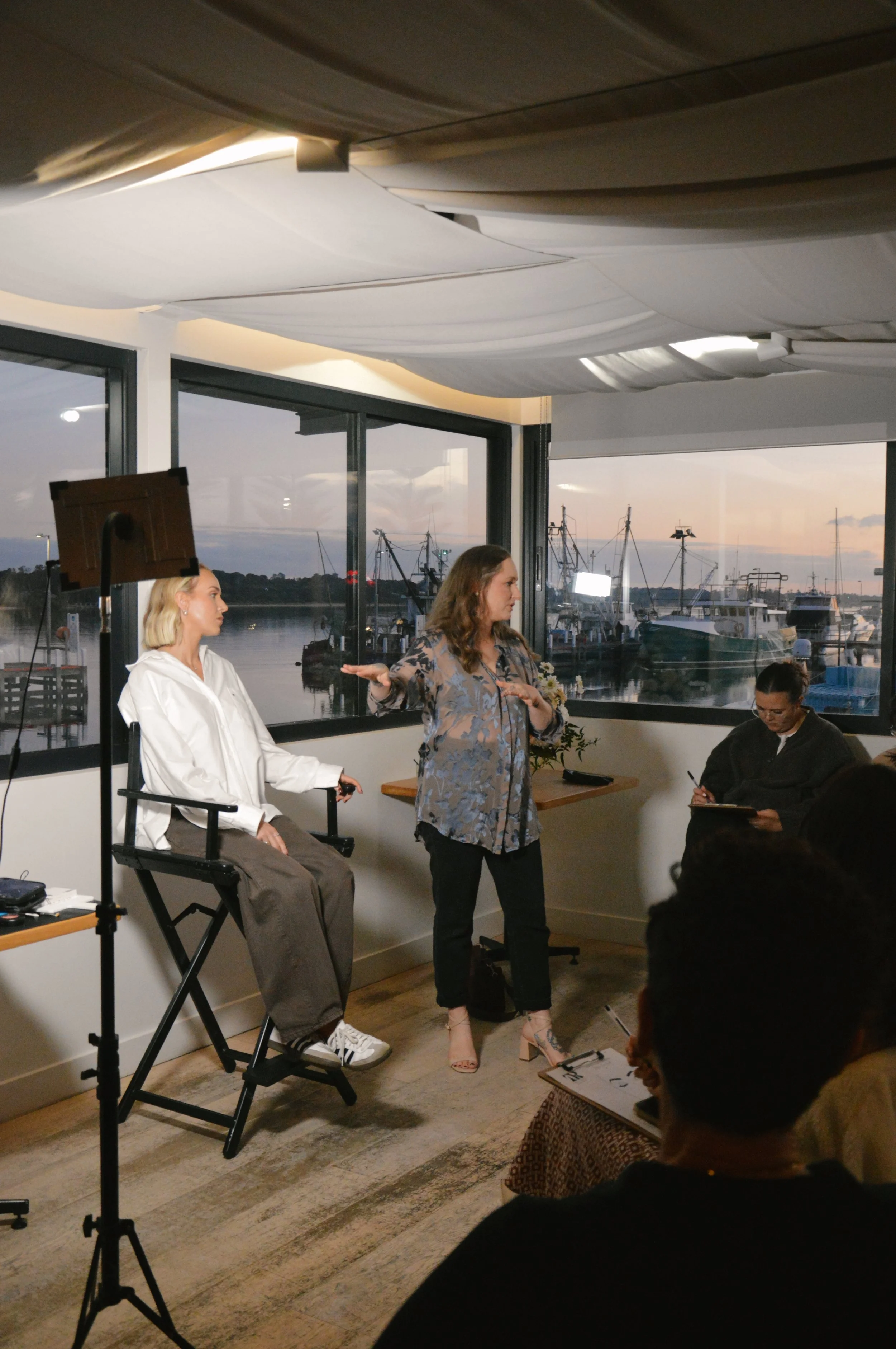 A woman standing and speaking to a group in a room with large windows overlooking a marina with boats at sunset. Two women are seated, one with a white hoodie and the other with glasses, engaged in different activities.