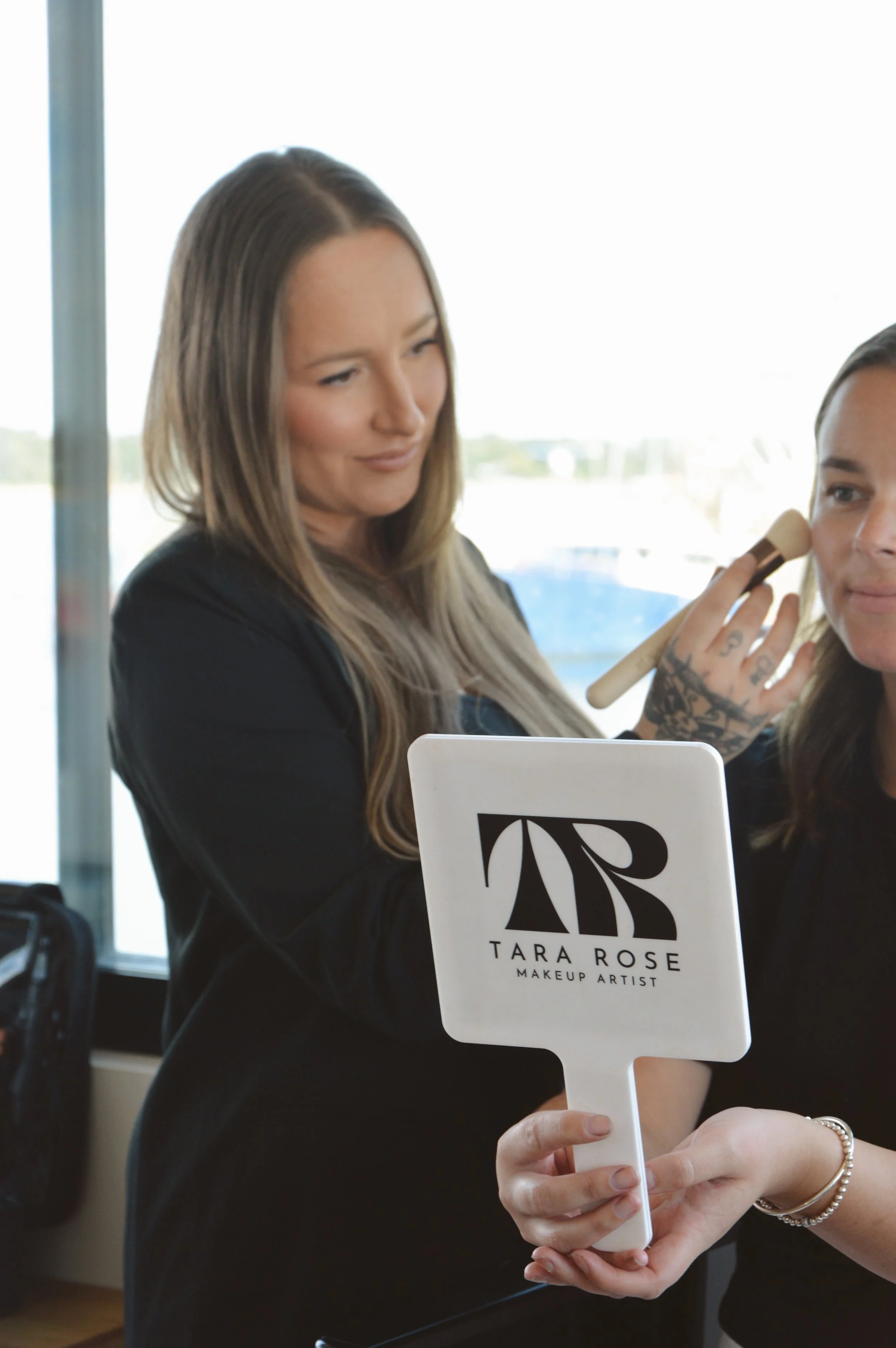 A makeup artist applying makeup to a woman in a well-lit room with large windows. The artist is holding a brush near the woman's face, and a sign with the name 'Tara Rose' and 'Makeup Artist' is visible in the foreground. The woman has long hair and is wearing a black outfit.