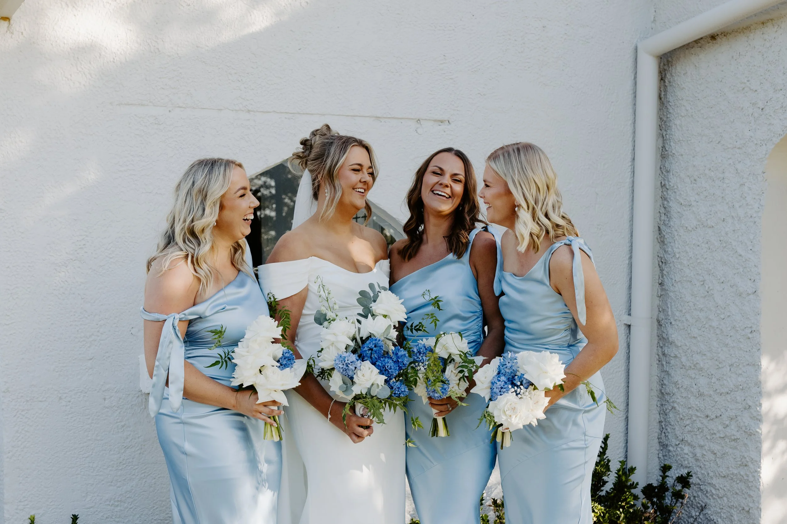 A group of five women, one in a wedding dress and four in matching light blue bridesmaid dresses, standing together outdoors against a white textured wall, all smiling and holding bouquets of white and blue flowers.