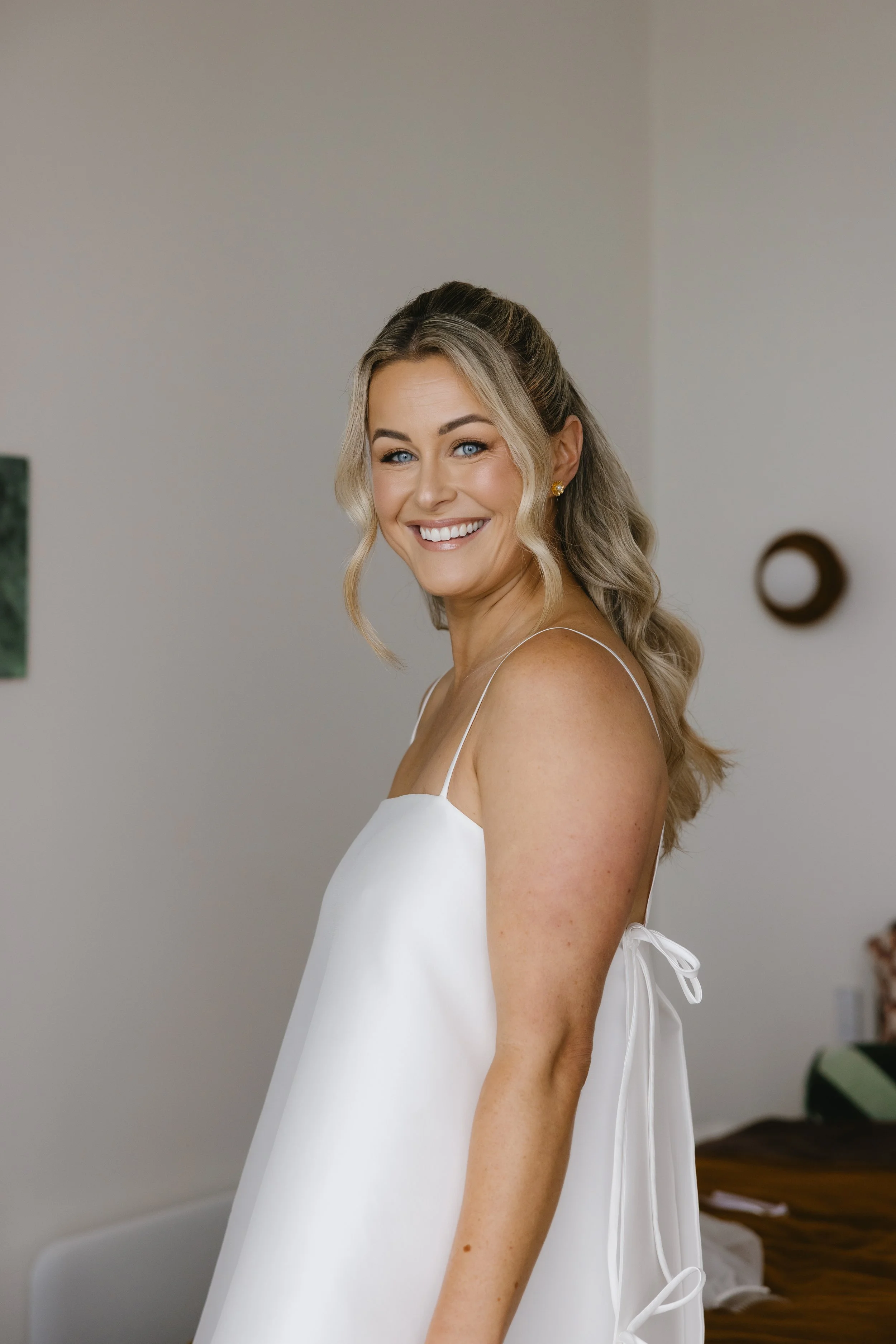 A smiling woman with blonde hair styled in loose waves, wearing a white dress with thin straps and a tied bow on her back, standing indoors against a neutral wall.