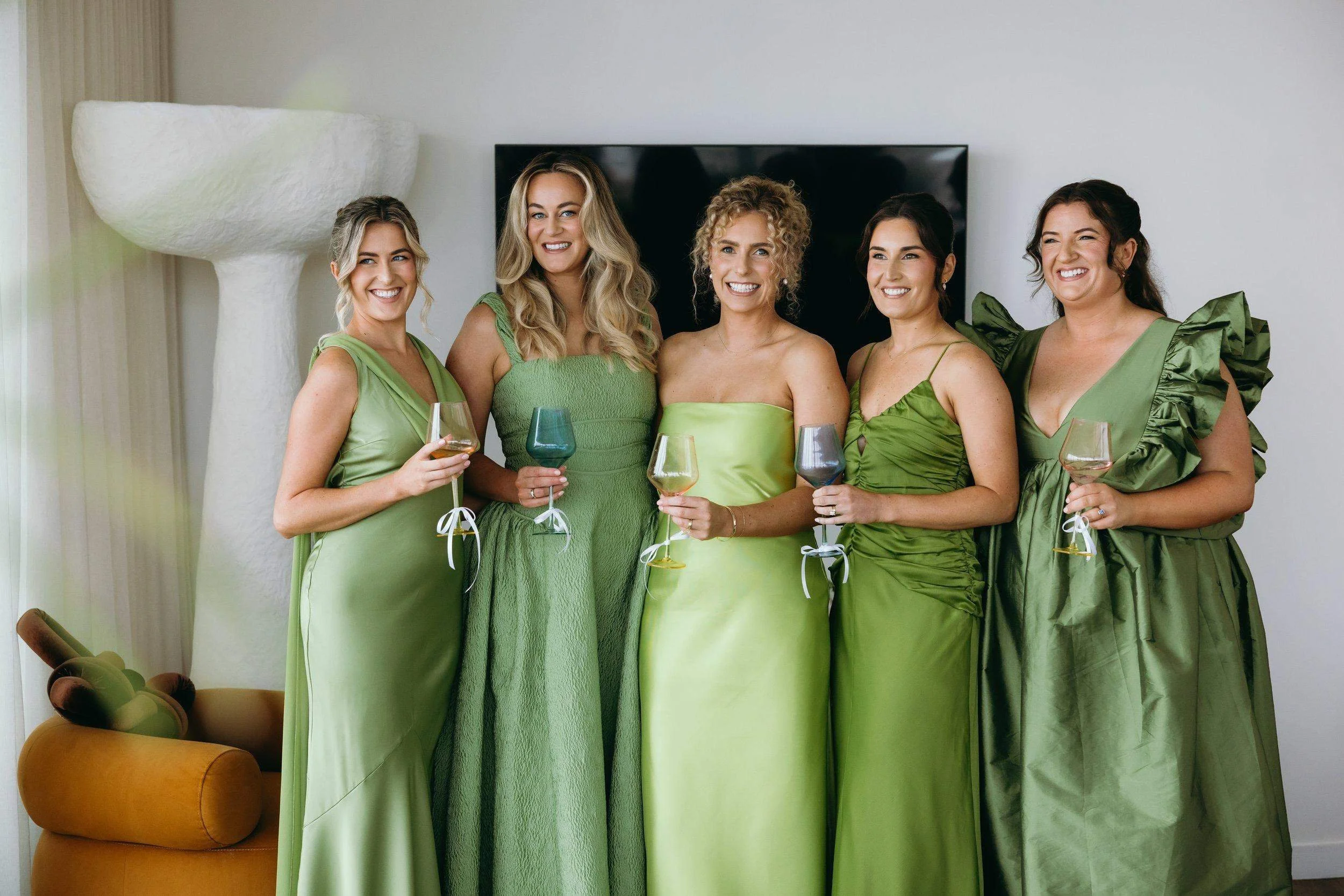 Five women dressed in various shades of green are smiling and holding wine glasses in a living room with a large white sculpture and a TV in the background.