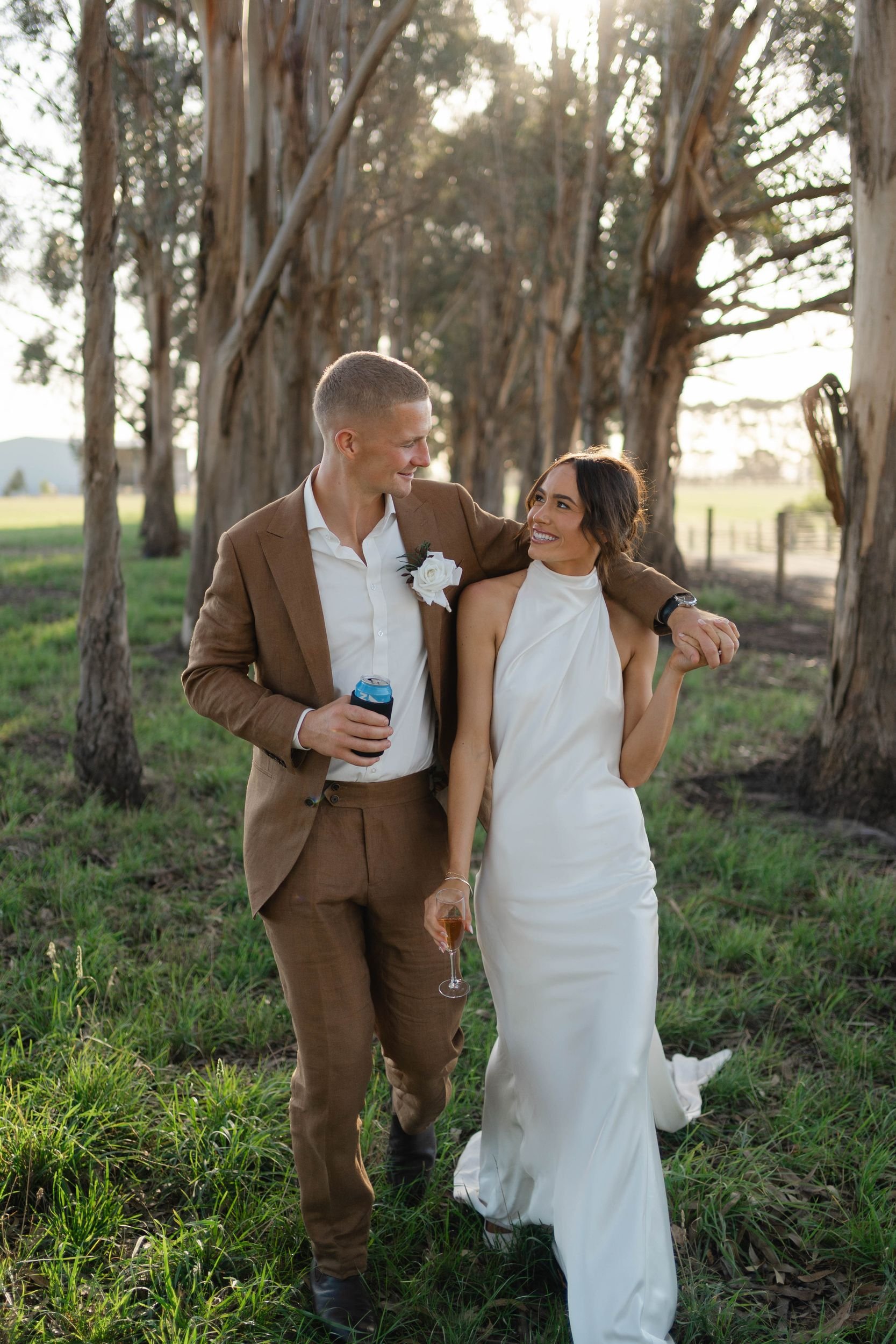 A man and a woman walk arm in arm through a grassy outdoor area with trees in the background. The man is wearing a brown suit and white shirt, holding a beverage can, and looking at the woman. The woman is wearing a white dress and holding a glass of