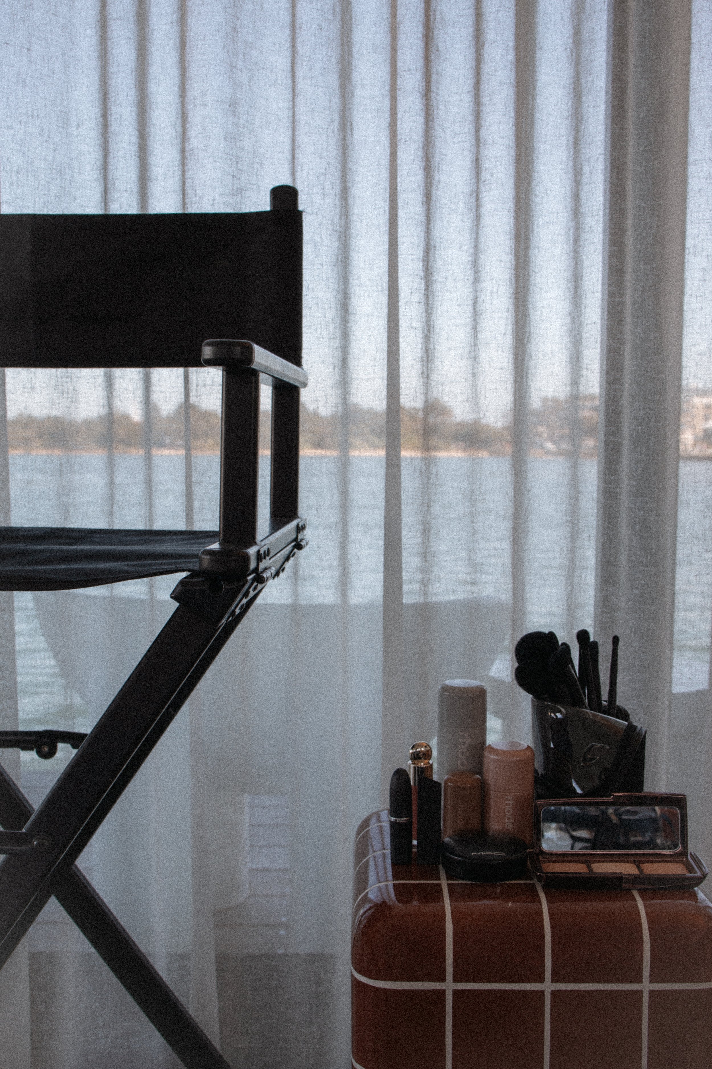 A makeup table with various cosmetic products, brushes, and a makeup palette, next to a chair and a window with sheer curtains.