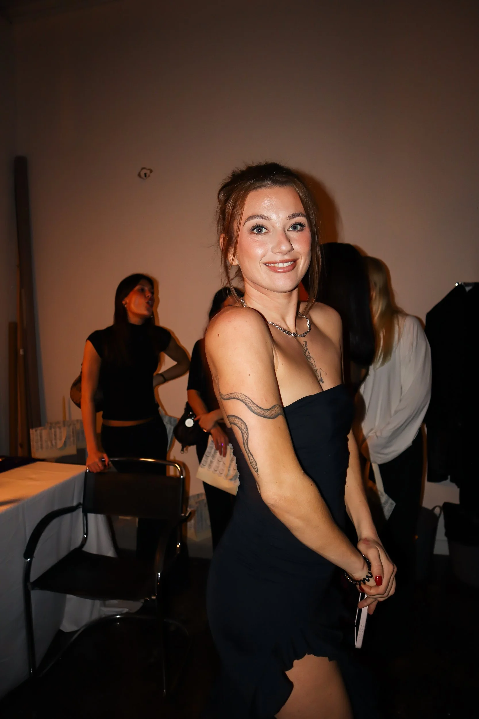 A woman with brown hair and tattoos on her arms in a black dress, smiling at the camera at a social event with other women in the background.