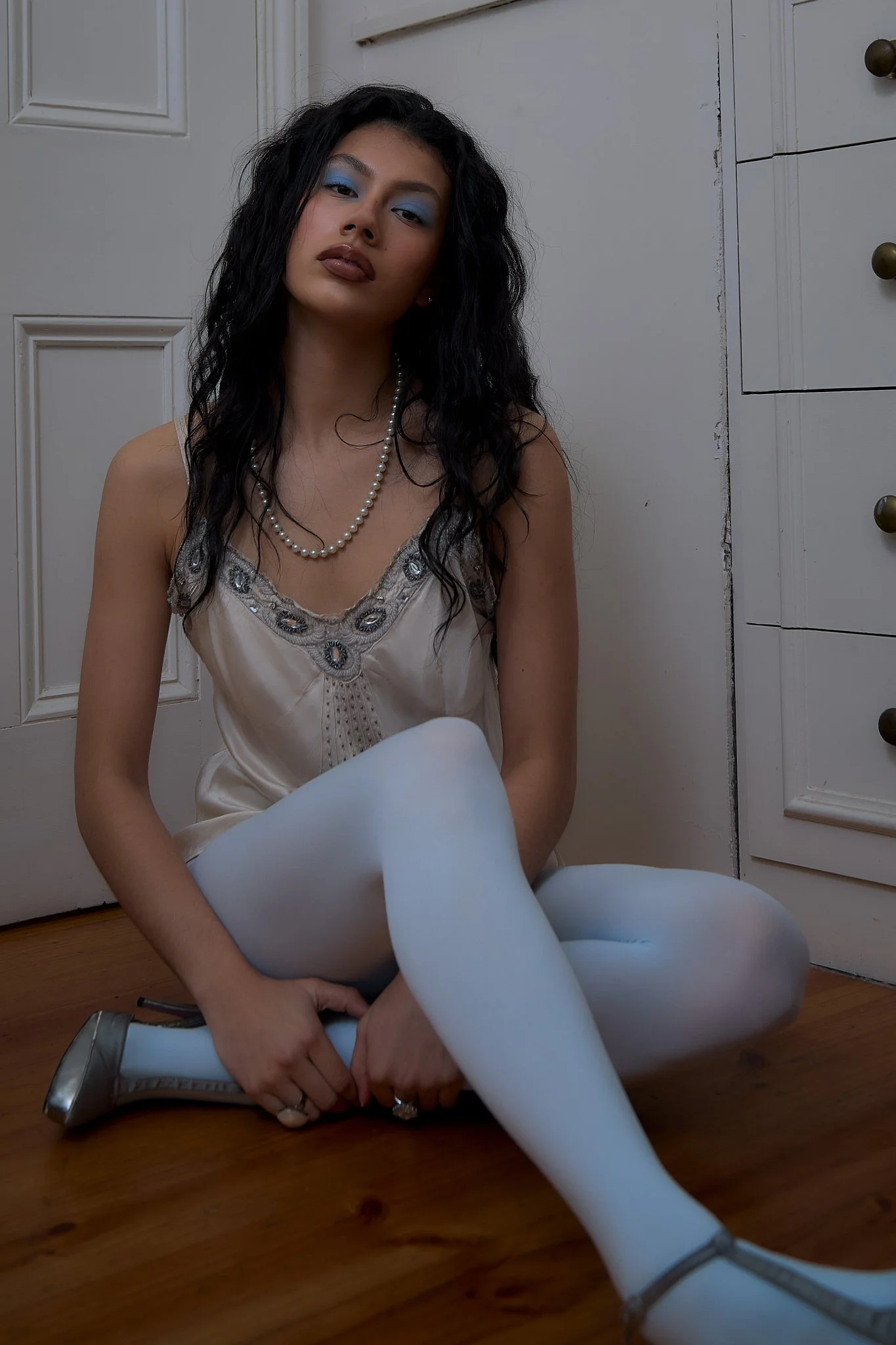 A young woman with dark curly hair, blue eyeshadow, and brown lipstick, sitting on the floor in front of white cabinets. She is wearing a light-colored satin top with embroidery, a pearl necklace, and white tights. Her left hand is holding her right ankle, and she is wearing silver heels.