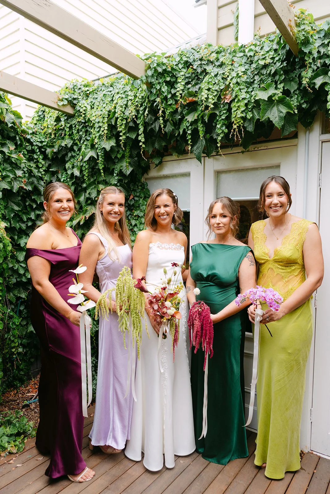 Five women dressed in colorful gowns standing together on a wooden deck, holding bouquets of flowers, with green ivy covering the wall behind them.
