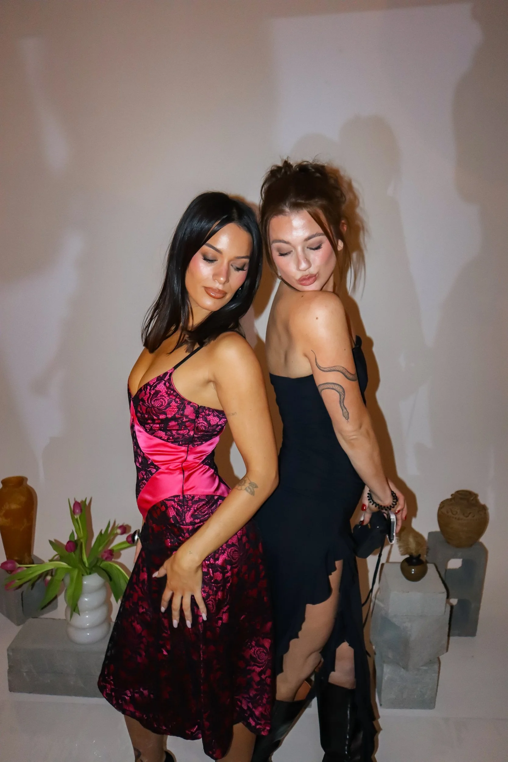 Two women with closed eyes and relaxed expressions, standing close together against a plain background. One woman has long black hair and wears a pink and black lace dress, while the other has red hair styled in a messy bun and wears a black dress with a snake tattoo on her arm. There are decorative vases and plants on a table behind them.