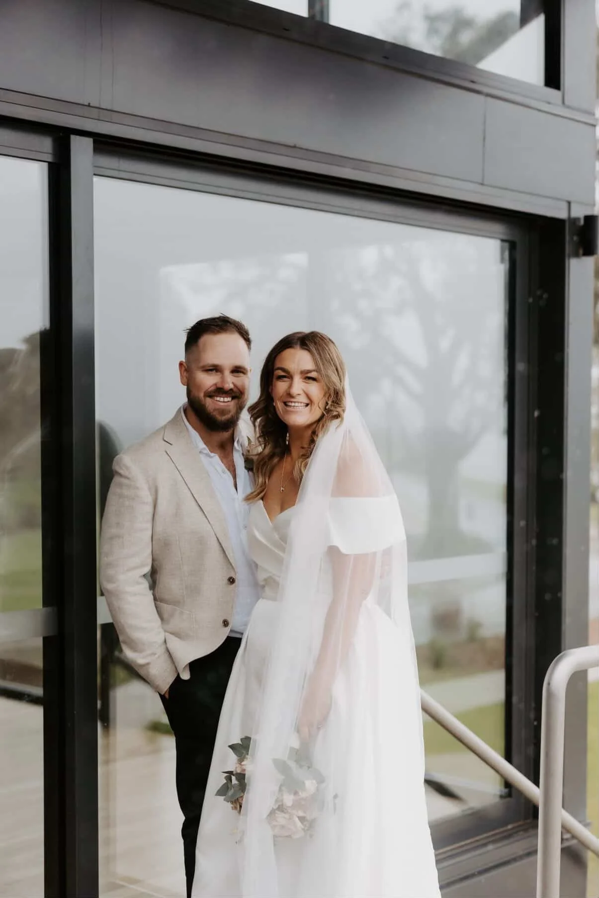 A newlywed couple in wedding attire standing together outside a glass building, smiling for the camera.