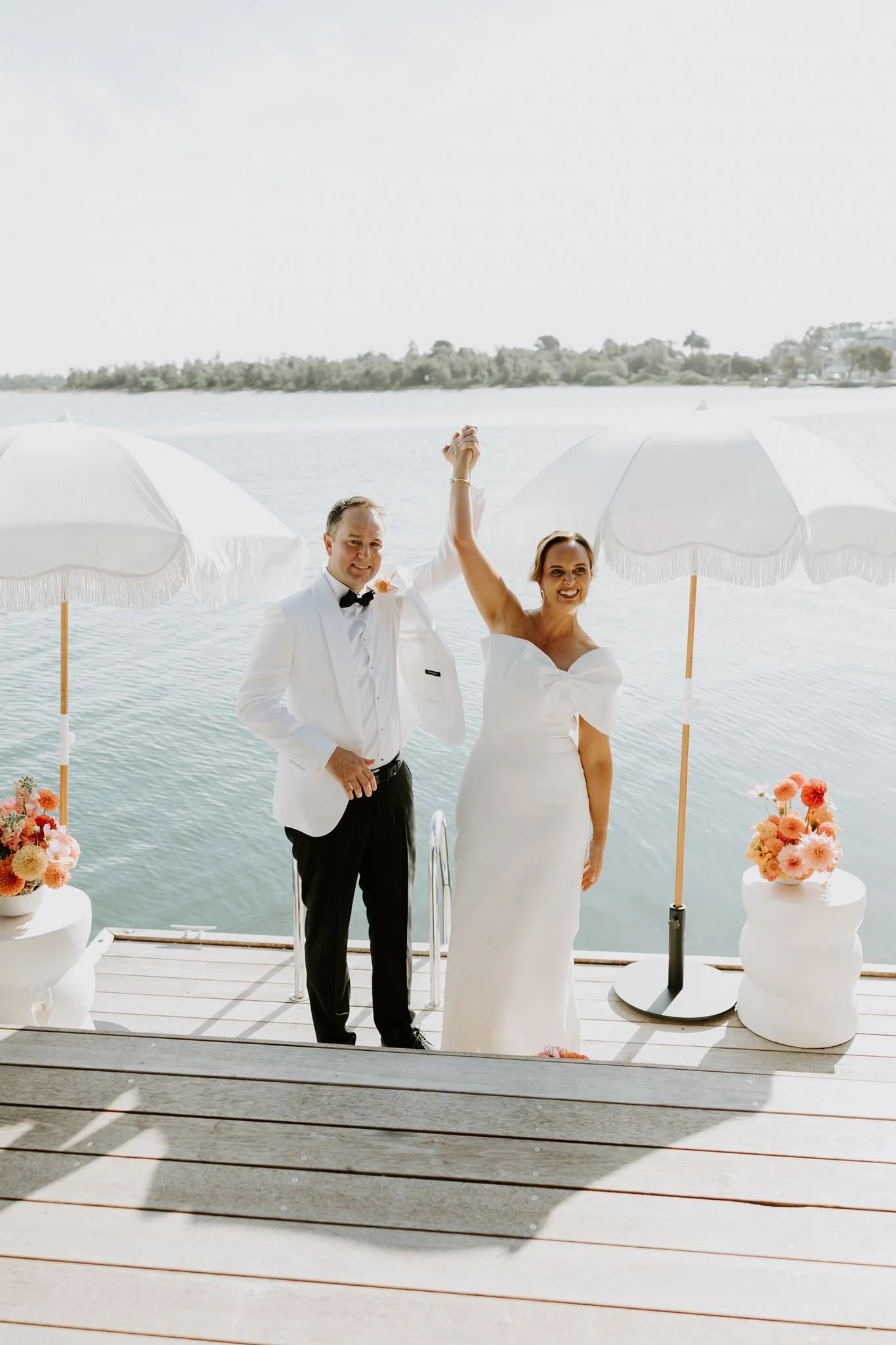 A newly married couple on a dock by a body of water, with the groom in a white tuxedo and the bride in a white wedding dress, holding hands and celebrating under white umbrellas and flower arrangements.