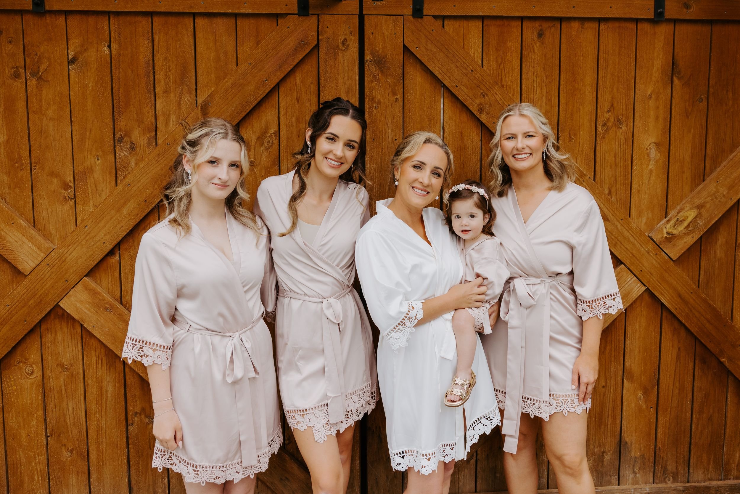 Group of five women, including a young girl, all dressed in white robes, standing in front of a wooden barn door, smiling.