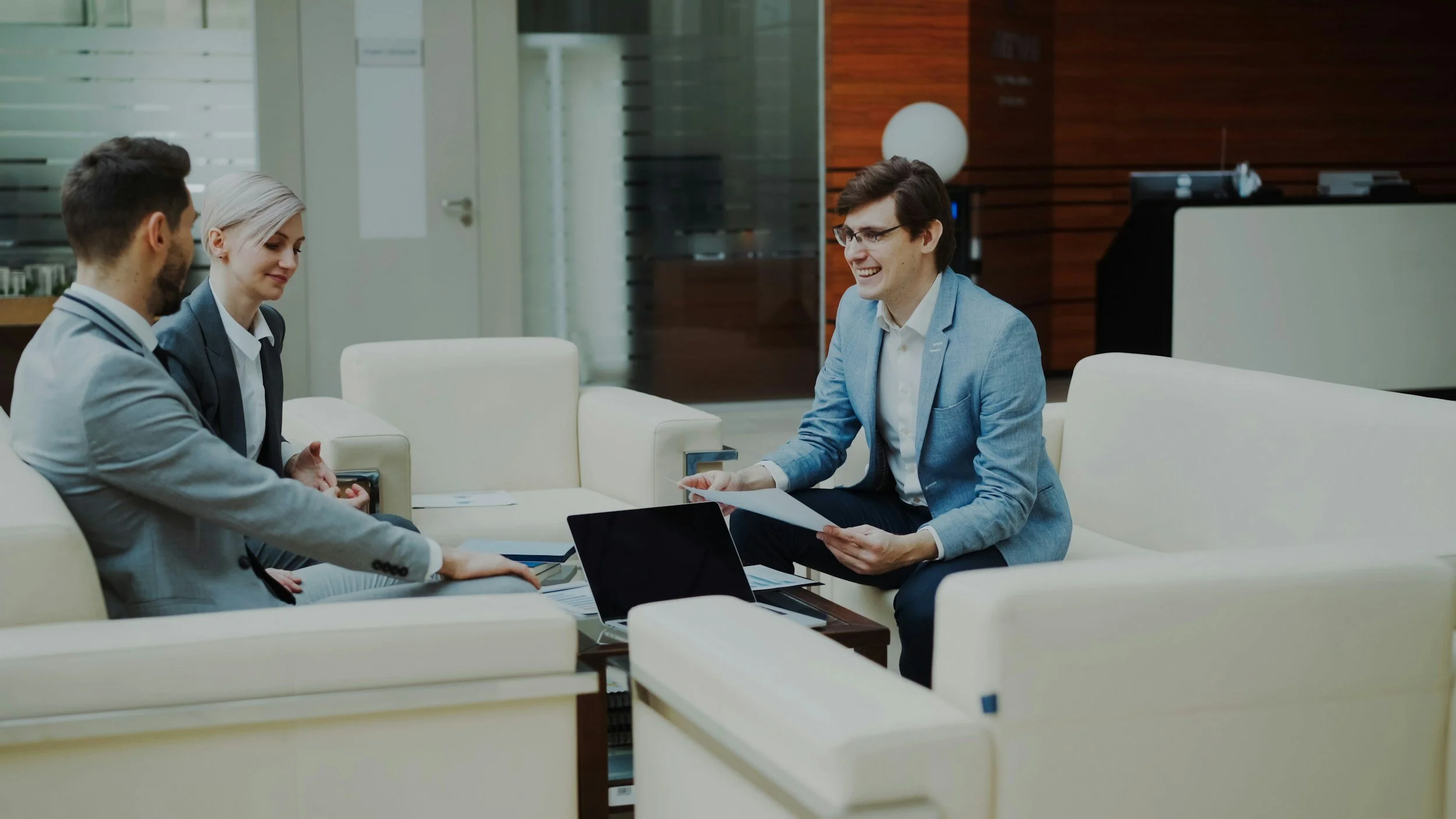 Three business professionals having a meeting in a modern office lounge with white sofas and a wooden accent wall, one man in a light gray suit, one woman in a navy suit, and another man in a blue blazer, all smiling and engaging in discussion.
