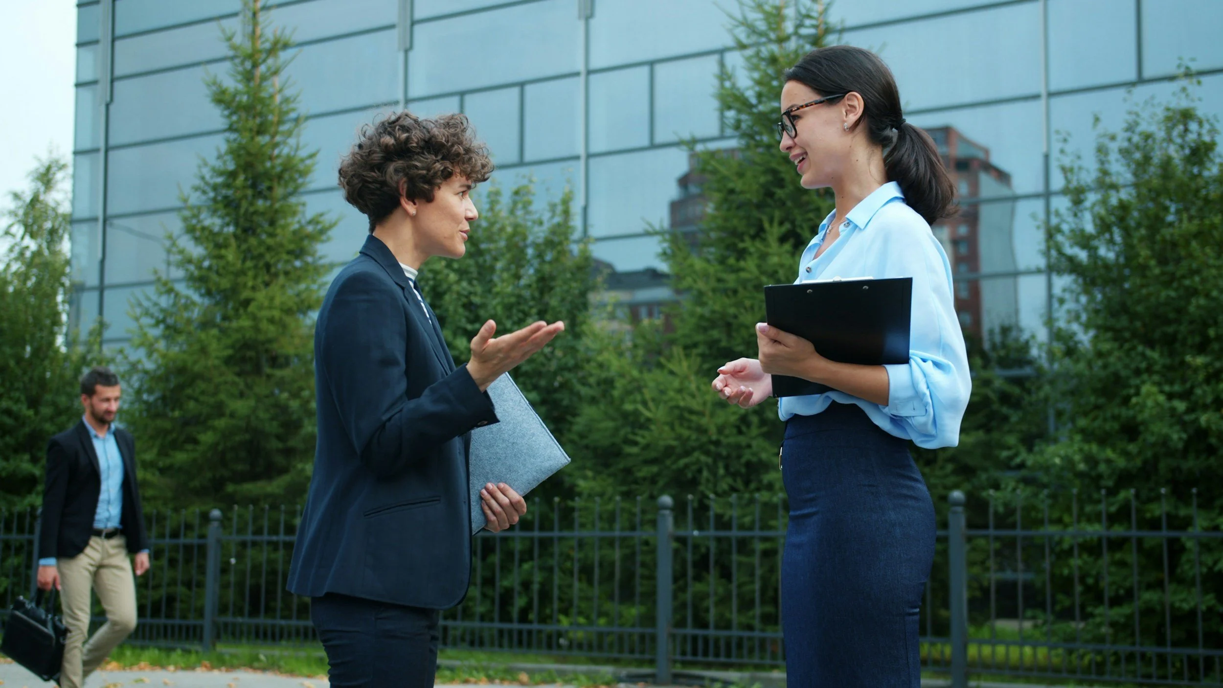 Two women having a conversation outdoors in front of a modern glass building and green trees. One woman has curly hair, wearing a dark blazer, and is holding a grey folder. The other woman has straight hair tied back, glasses, and is wearing a light blue blouse and dark skirt, holding a black folder. A man is walking by in the background.