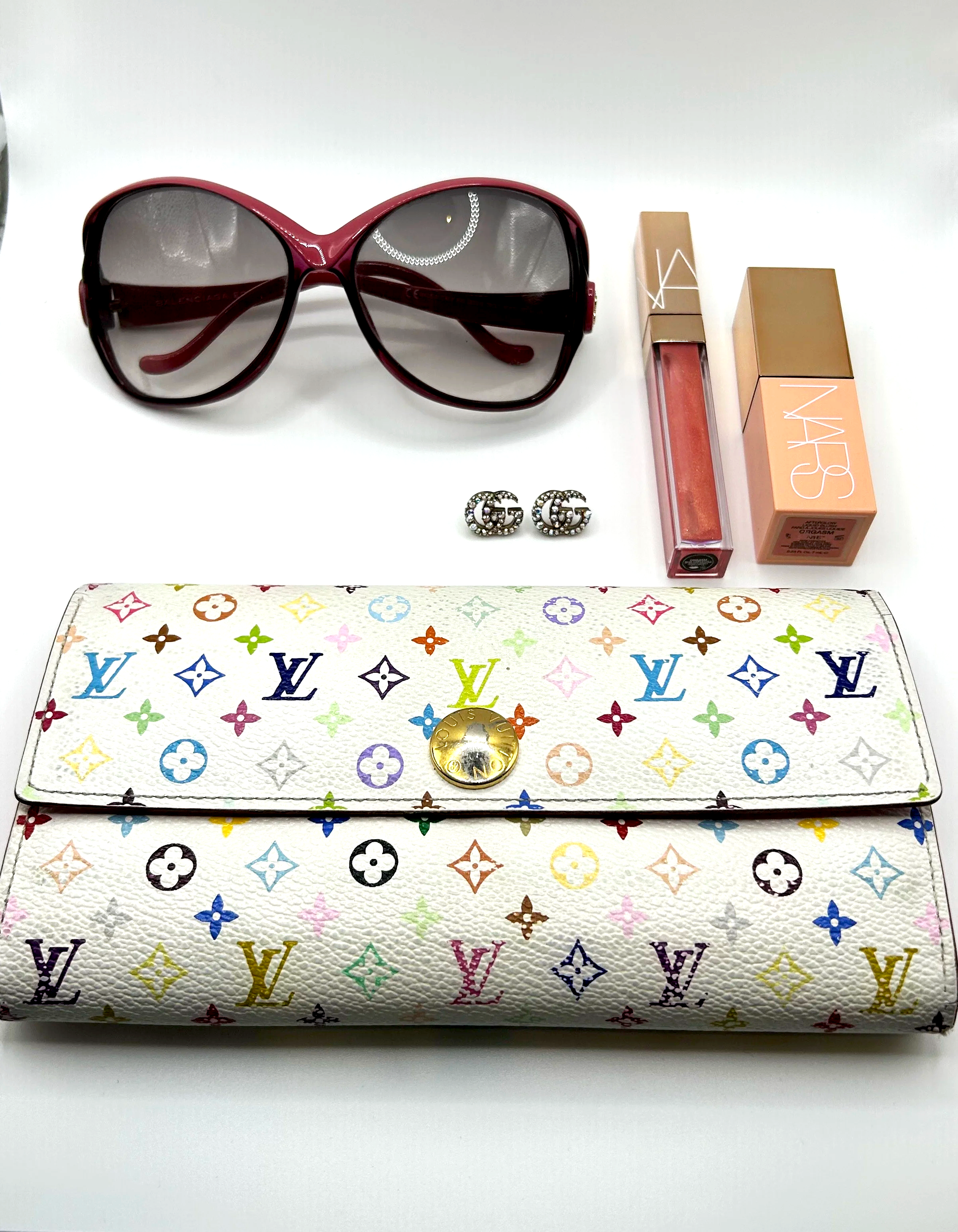 A pair of large red sunglasses, two pairs of GG earrings, a NARS lip gloss, a NARS blush, and a multicolored Louis Vuitton wallet on a white surface.