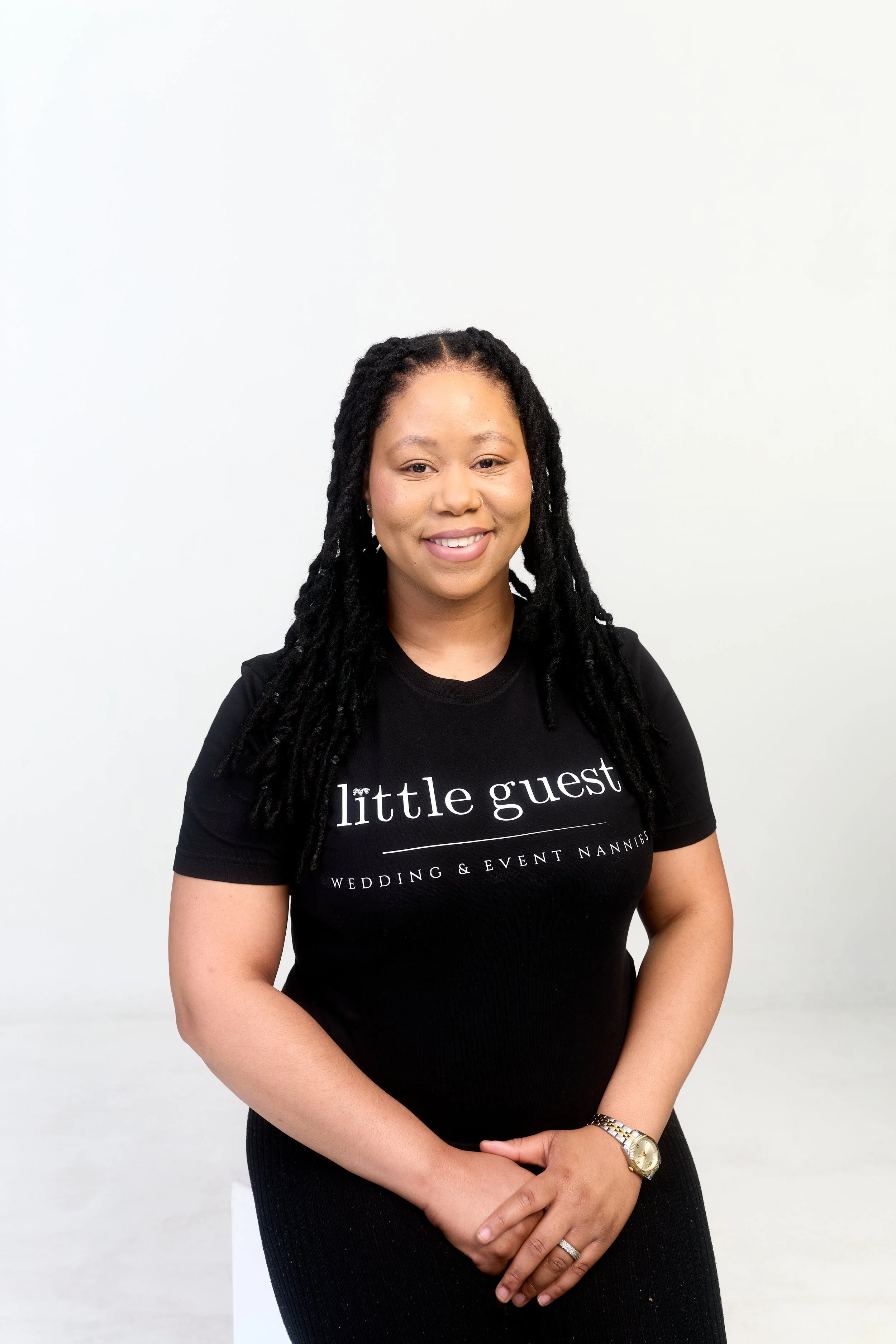 A woman with long black hair in dreadlocks, wearing a black T-shirt with white text that reads 'little guest Wedding & Event Nannies,' standing against a plain white background, smiling with her hands clasped in front of her, wearing a gold watch and ring.