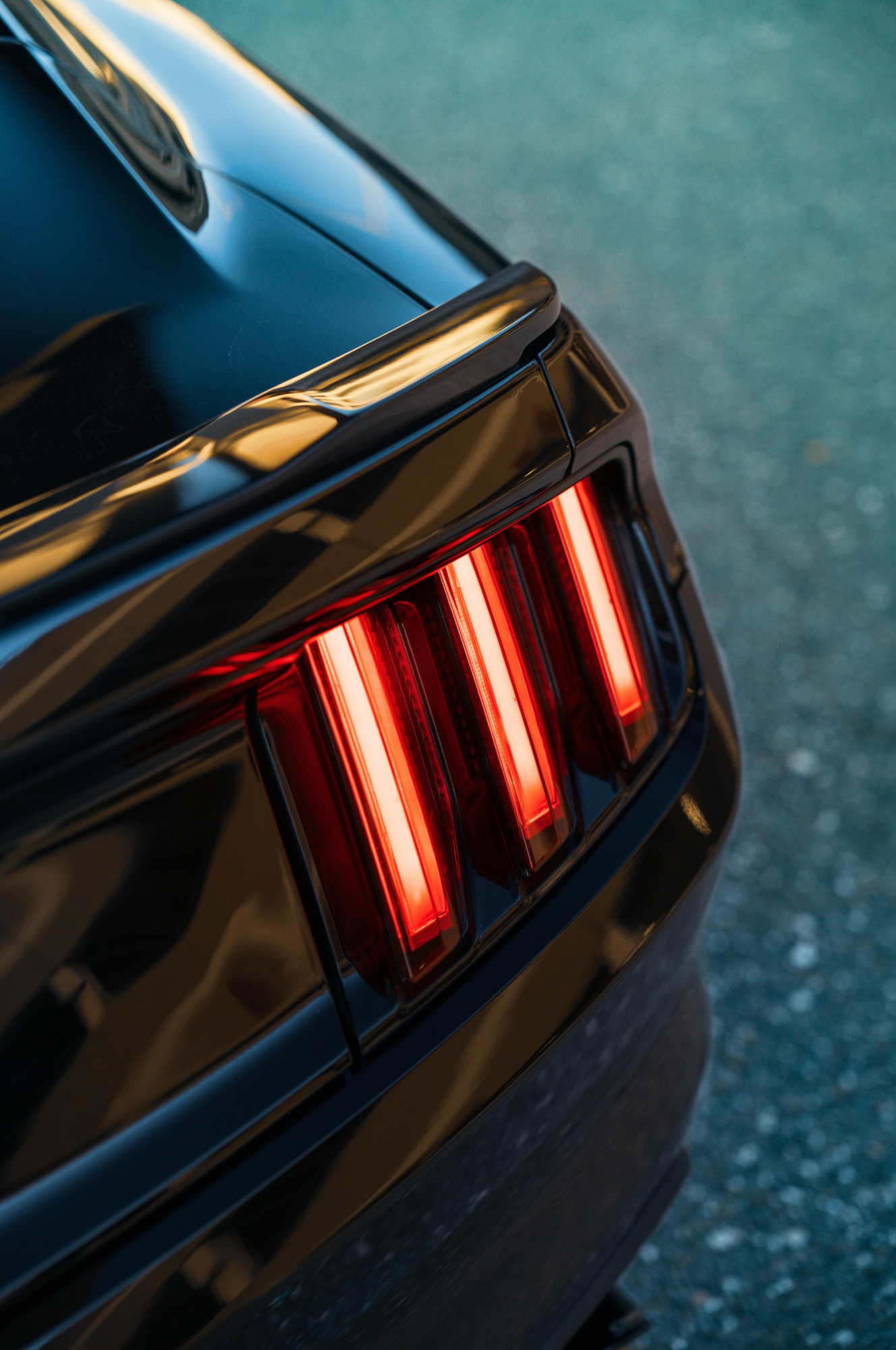Close-up of a black car's rear with illuminated vertical taillights and a sleek, shiny surface, on paved ground.