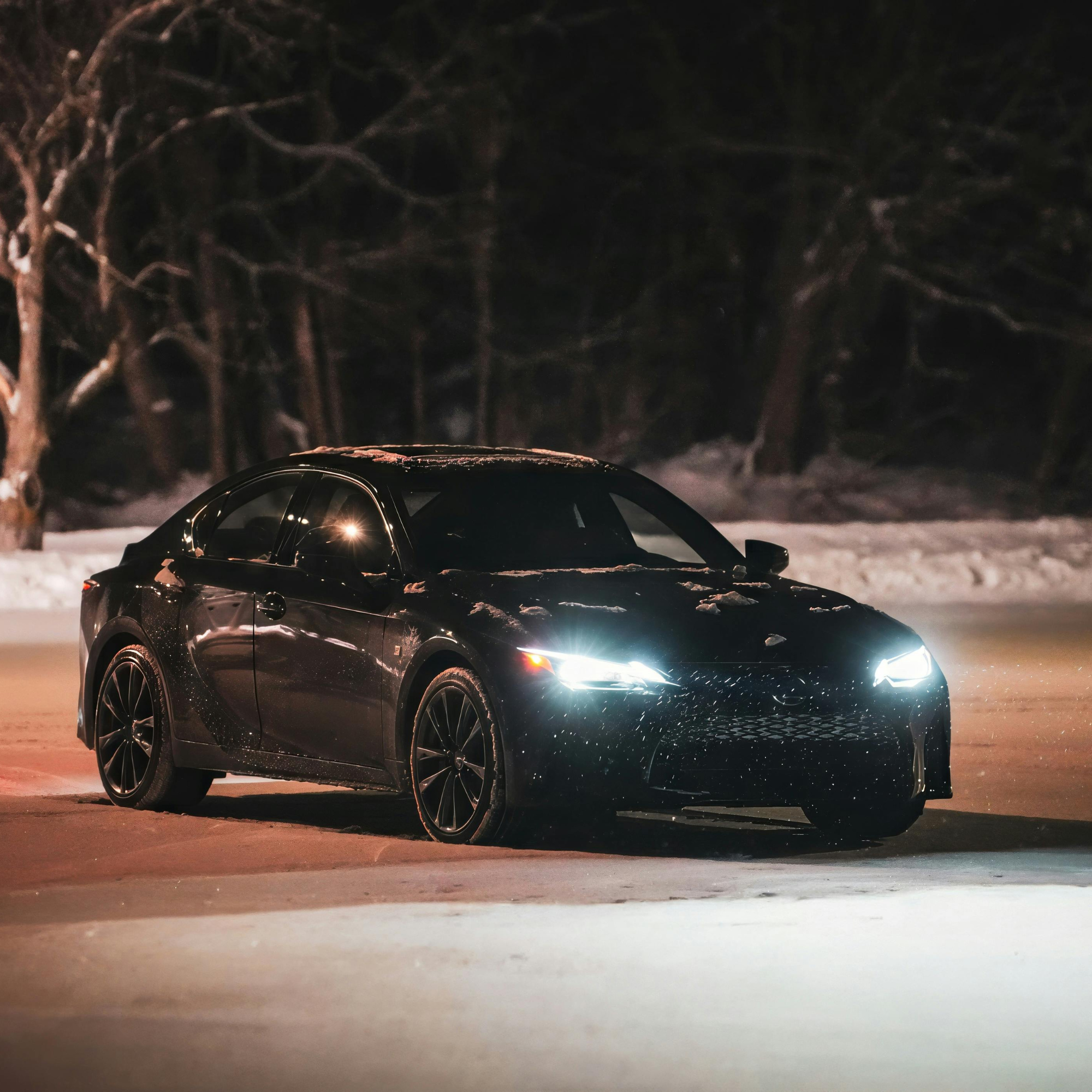 A black car parked outdoors at night on snow-covered ground, with snow on its roof and a snowy background of trees, illuminated by beam headlights.