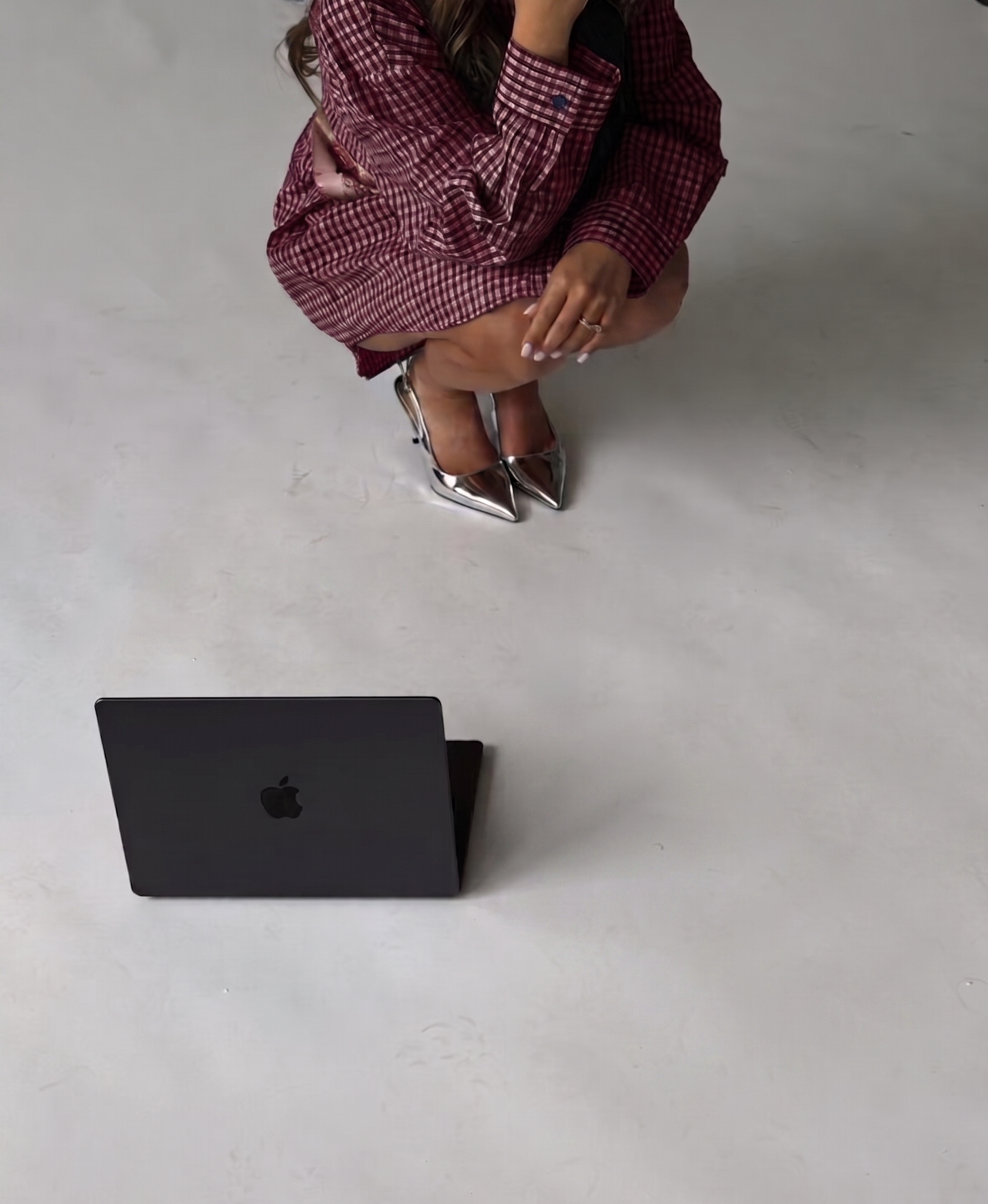 A woman in a red checkered dress with silver high heels, squatting indoors on a white floor near a black Apple MacBook laptop.