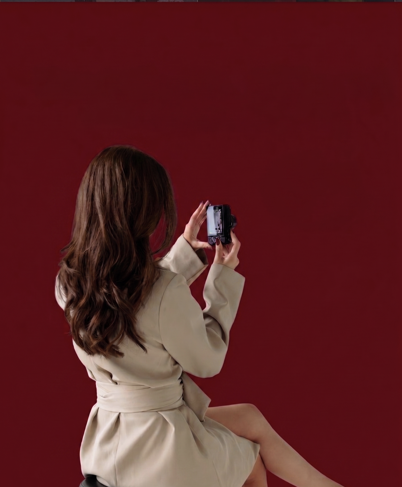 Woman with brown hair wearing a beige coat sitting and taking a photo with her camera against a red background.