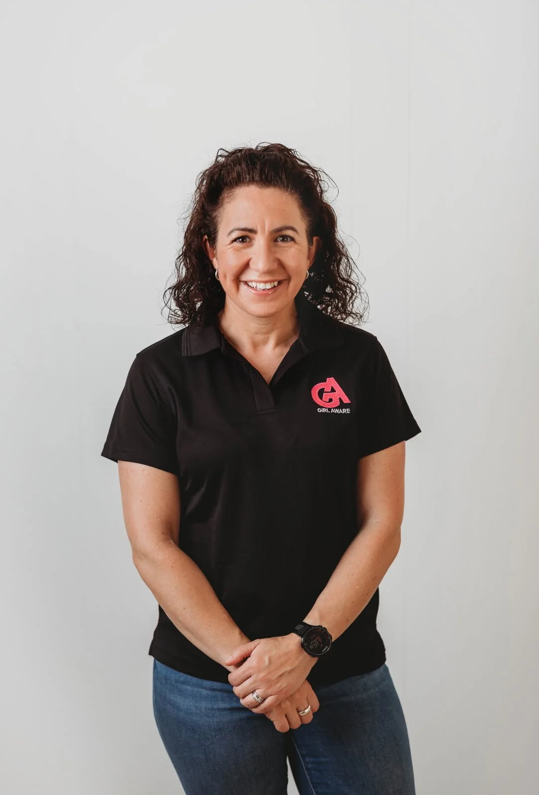 A woman standing against a plain gray background, smiling, wearing a black polo shirt with a pink and white logo that reads 'Girl Aware,' and blue jeans.