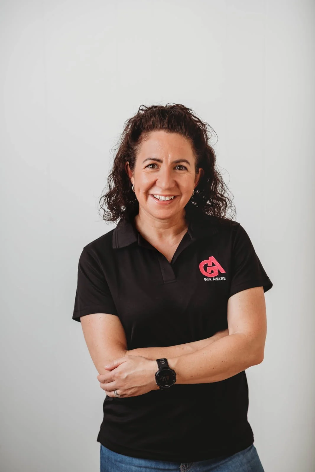 A smiling woman with dark, curly hair, wearing a black polo shirt with a pink and white logo that says 'GIRL AWARE,' standing against a plain white background.